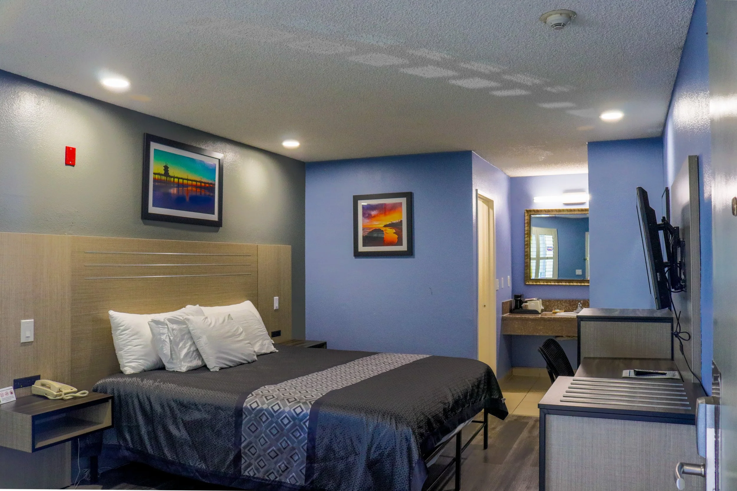 Interior of a hotel room with a bed, two framed landscape pictures on the wall, a small bedside table with a telephone, and a desk with a mounted TV. There is a window with shutters and a mirror on the wall. The room has blue and gray walls, and the ceiling has four recessed lights.