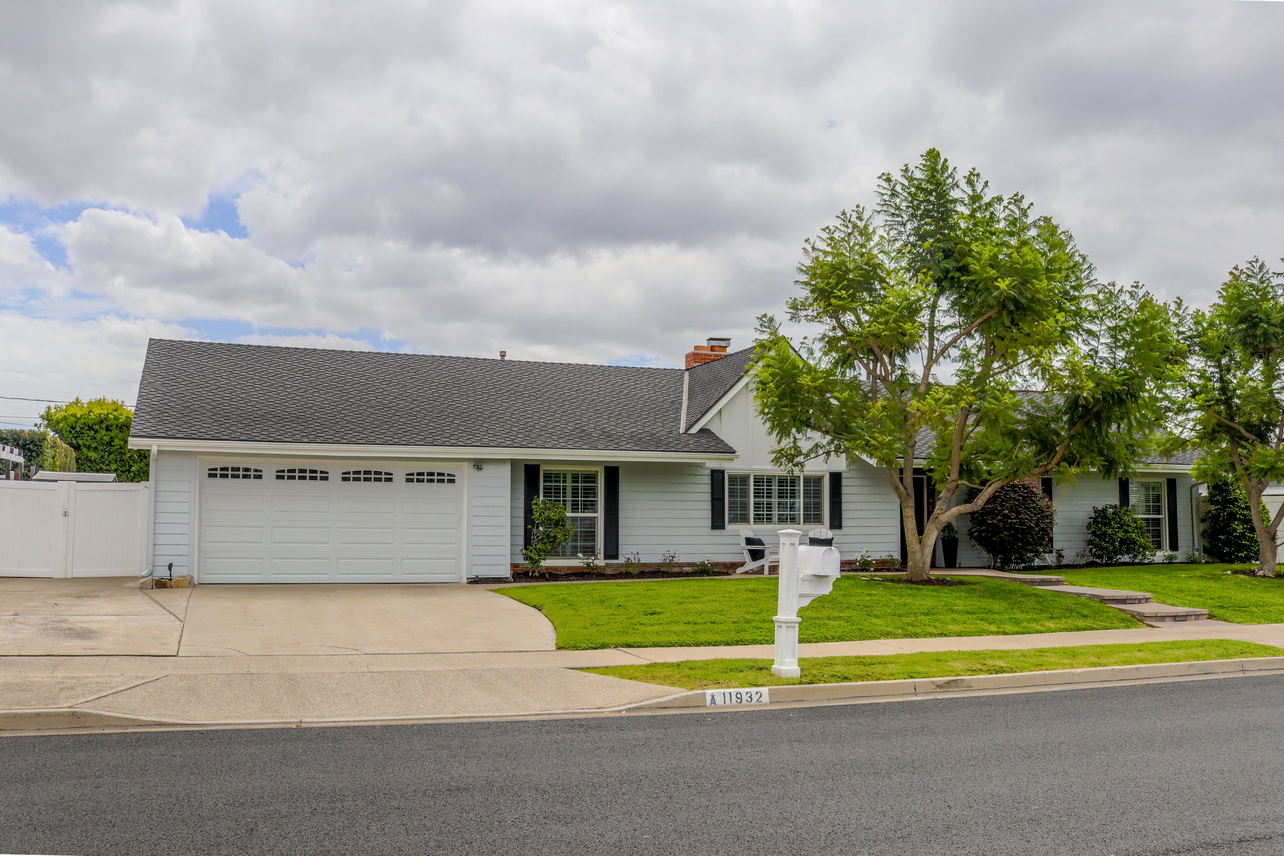 A suburban house with a gray shingle roof, white siding, and black window shutters, with a garage door showing. There are small front yard trees and bushes, a mailbox, and a concrete sidewalk in front. The street reads '11932' on the curb. The sky ab