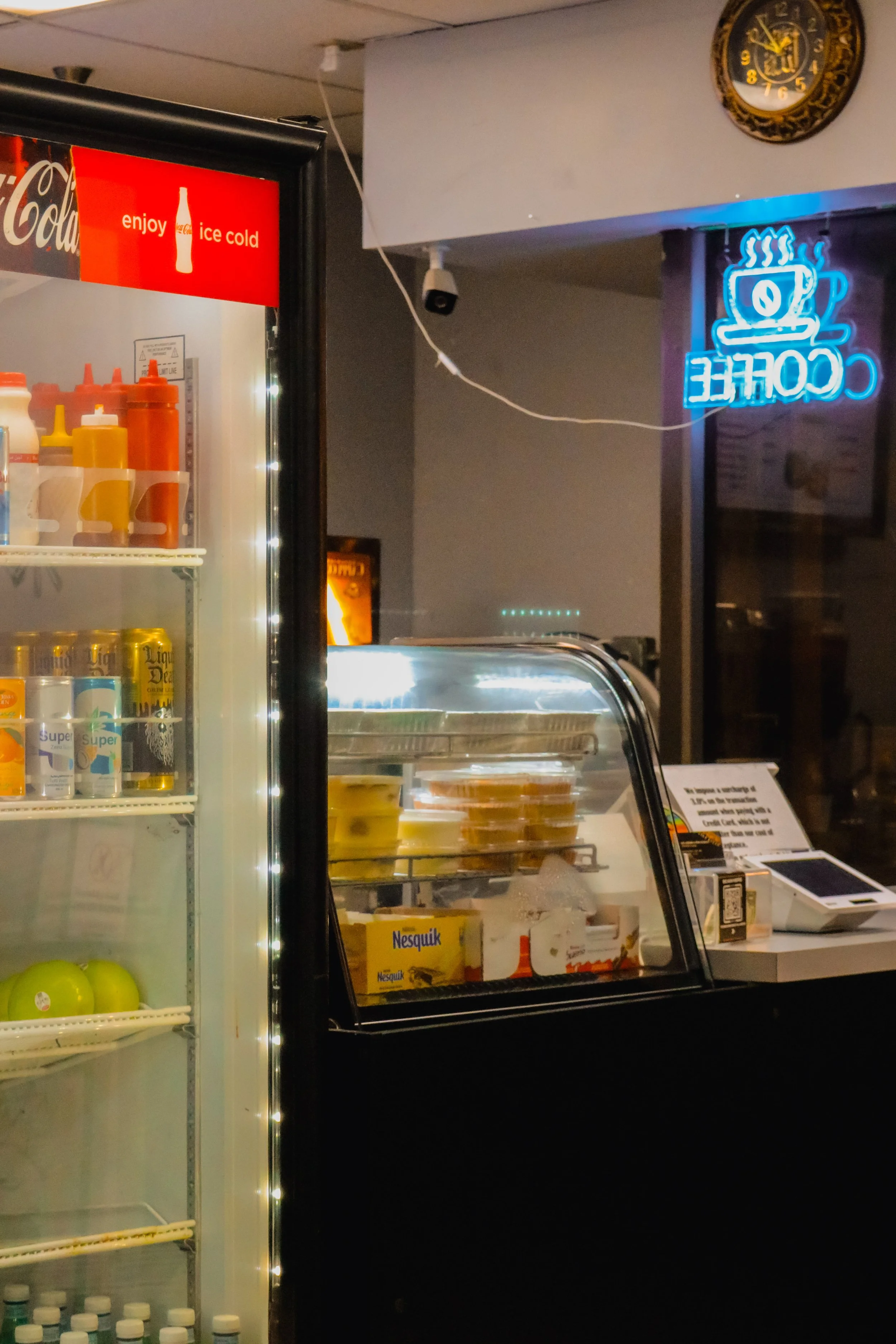 A refrigerated display case with food items inside, a chalkboard with the word 'COFFEE' and an illustrated coffee cup in neon blue, a sign that says 'enjoy ice cold', and a clock showing 11:54.