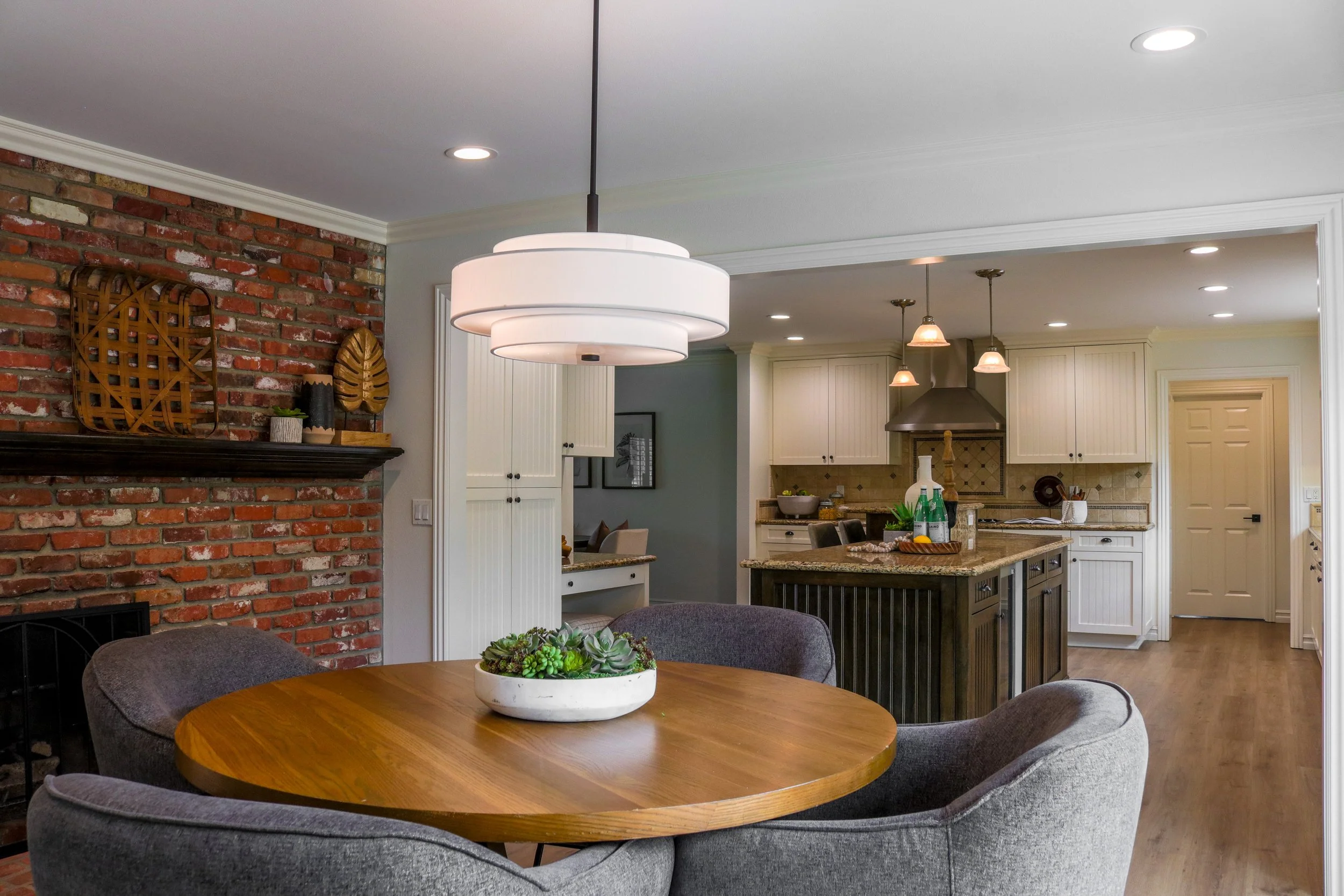 Living room with a red brick fireplace, modern white chandelier, and a wooden dining table surrounded by gray upholstered chairs. The kitchen with white cabinets, recessed lighting, and a central island is visible in the background.