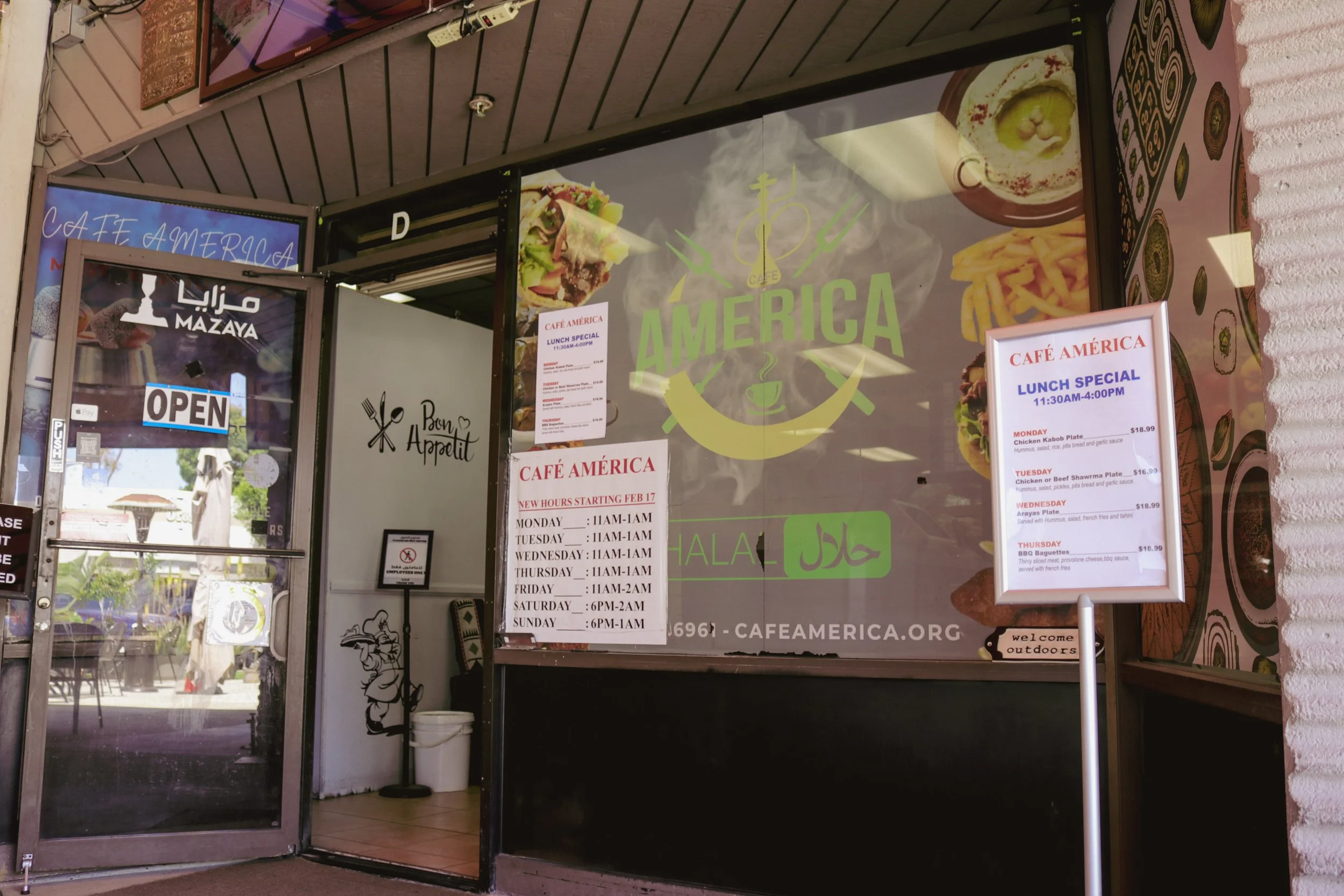 The image shows the entrance of Café América, a restaurant advertising a lunch special menu with various dishes listed. There are signs indicating that the café is open, its hours of operation, and signage in various languages including English and A