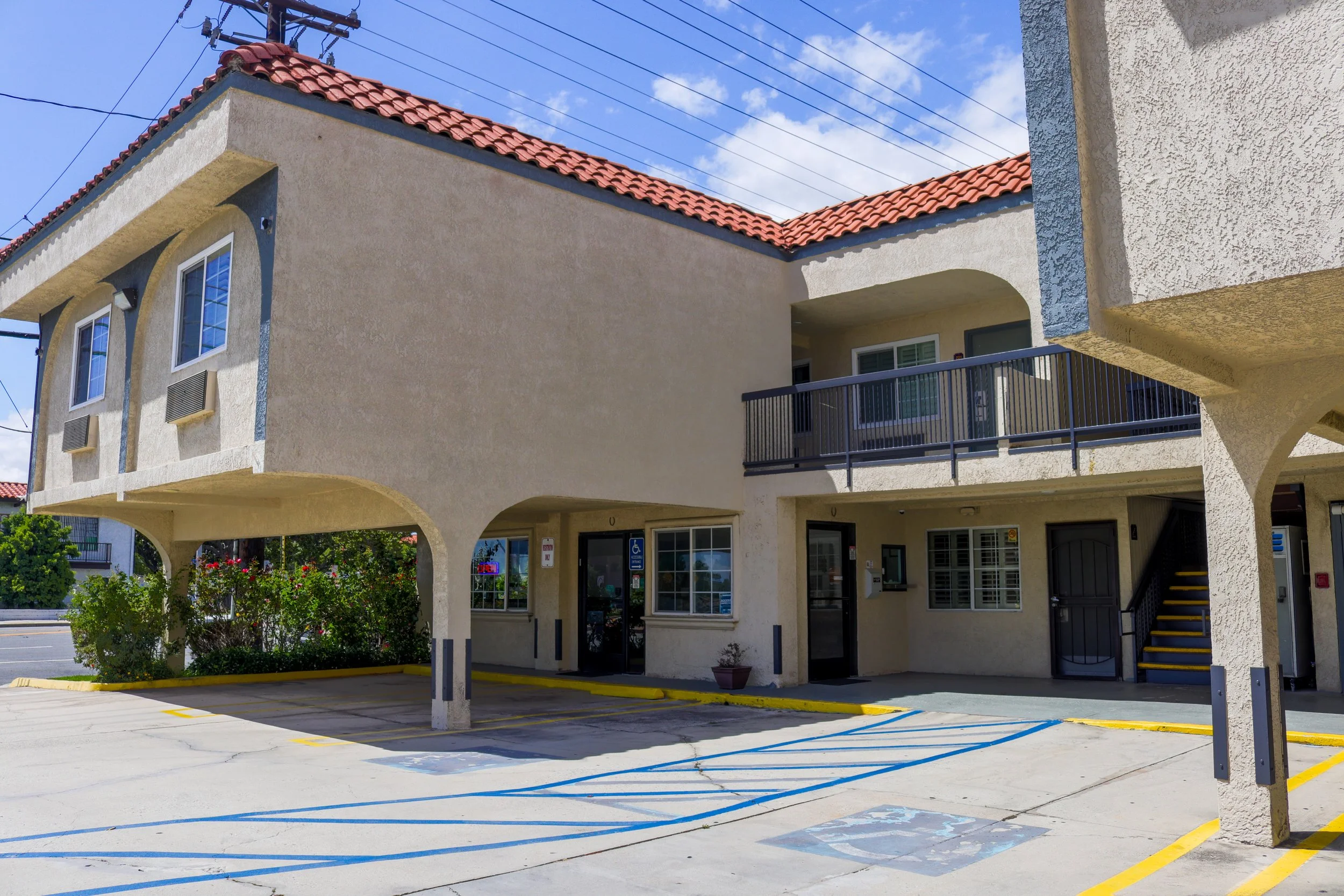 The image depicts a two-story beige residential building with a red tile roof, balconies, and multiple windows. In front, there's a parking area with painted parking spaces, and the building has an arched driveway. There are other buildings and some 