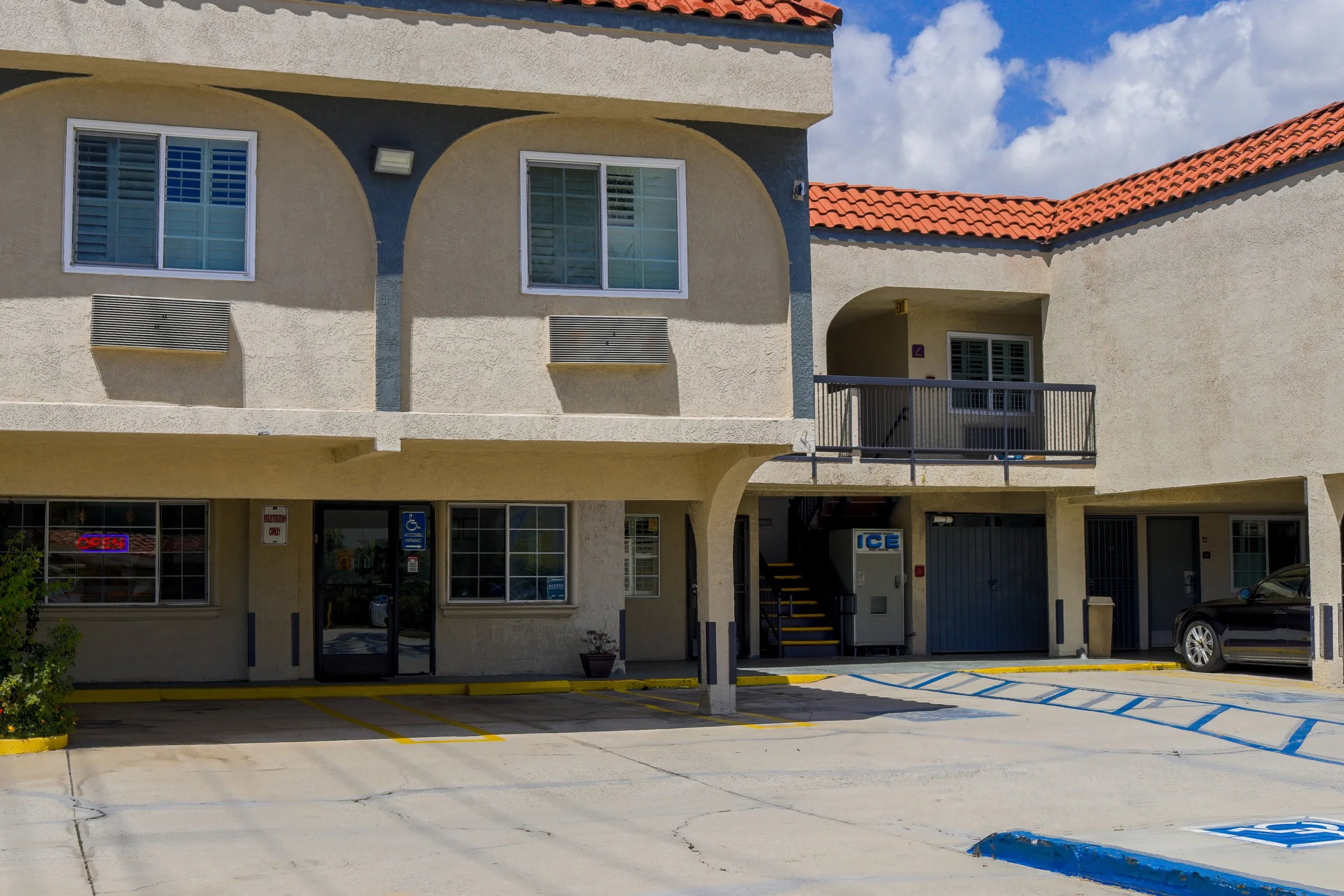 A multi-story building with beige walls, red tile roof, and various windows and vents. The ground level shows parking spaces, one with a car, and signs indicating handicapped parking and no parking zones. The upper level has balconies and windows wit