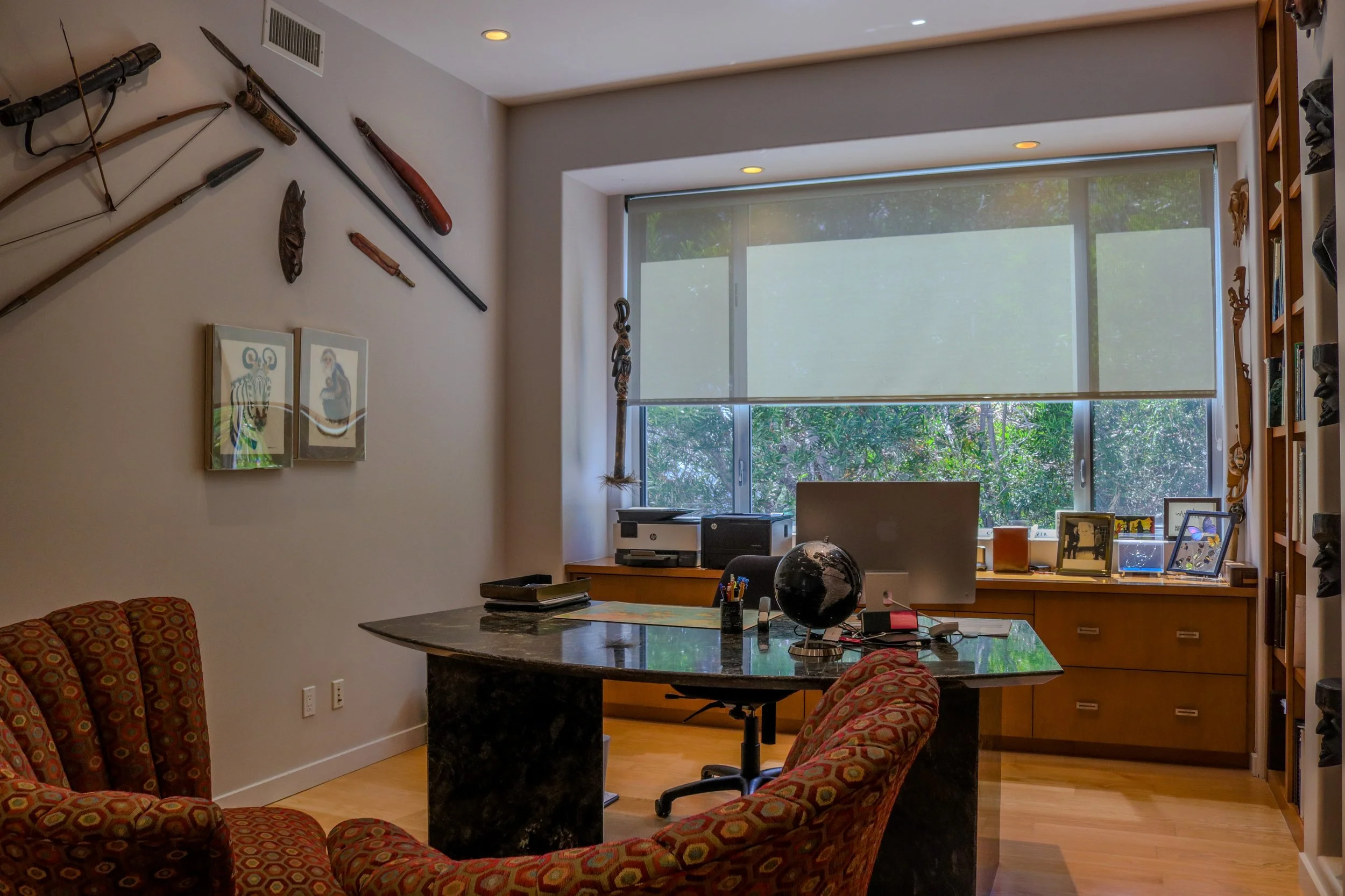 Interior of an office with a large window, desk, and chairs. The wall has traditional weapons, masks, and artwork displayed.