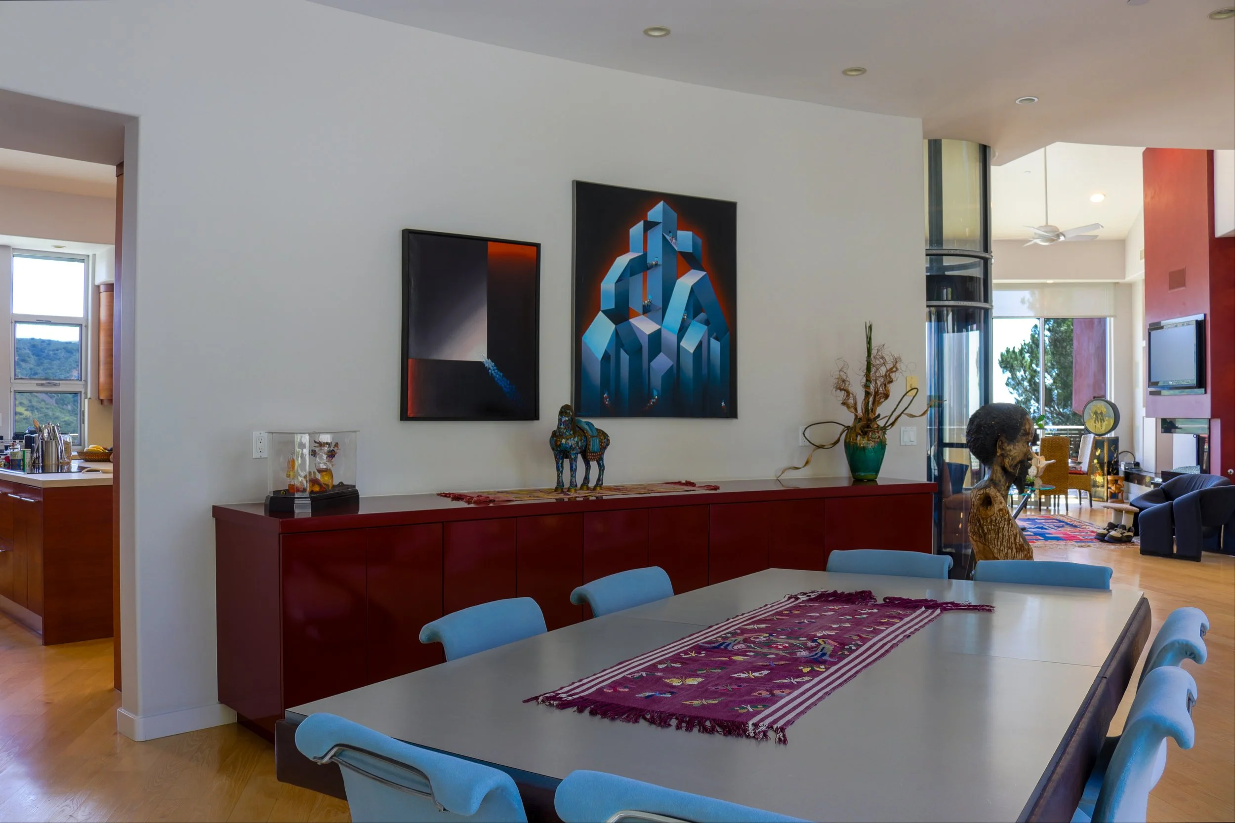 Living room with modern art on the wall, a red sideboard, sculptures, and large windows with a view of trees and hills.
