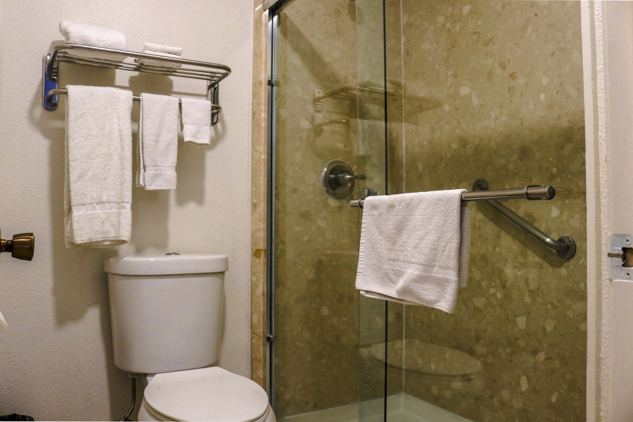 A bathroom with a toilet and a glass shower enclosure. The toilet has a shelf above it with three rolled white towels. A white hand towel hangs on a bar attached to the shower door. The shower has beige marble tiles.