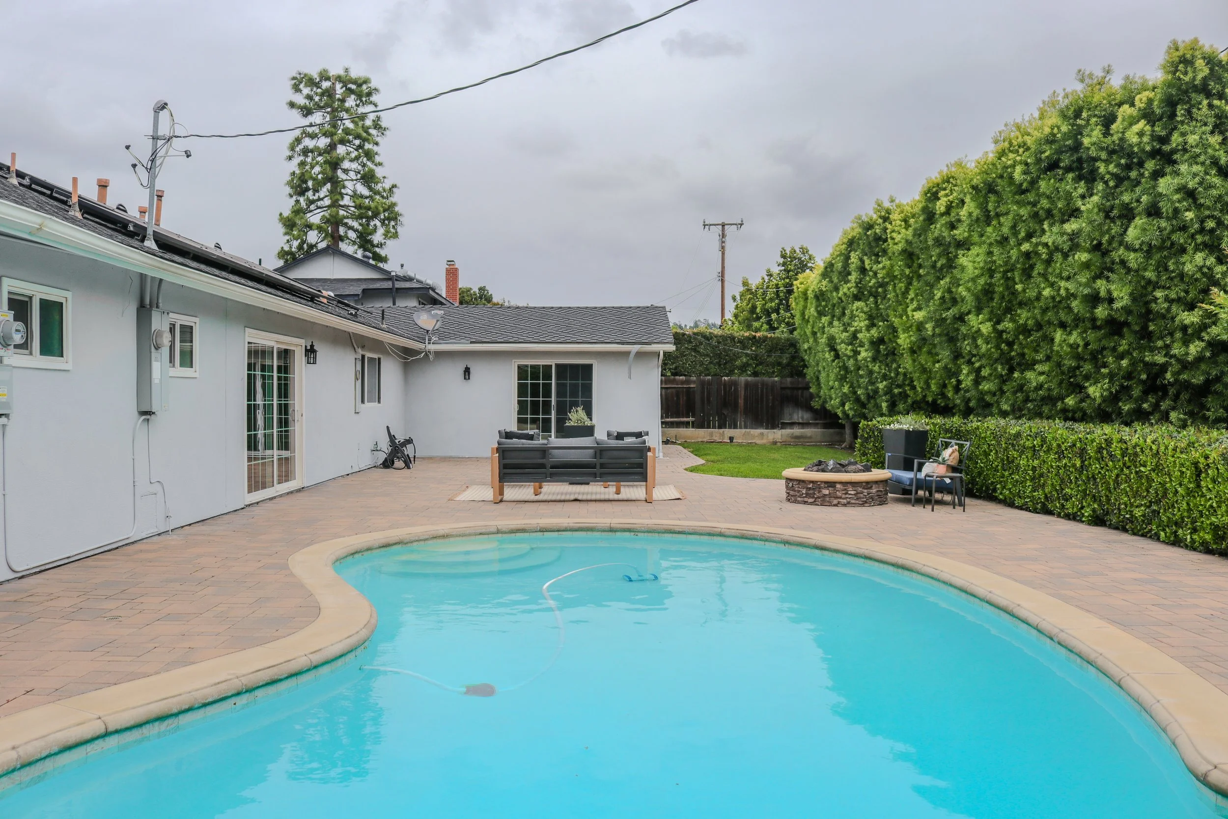 Backyard with a swimming pool, patio furniture, a large green tree, and a cloudy sky.