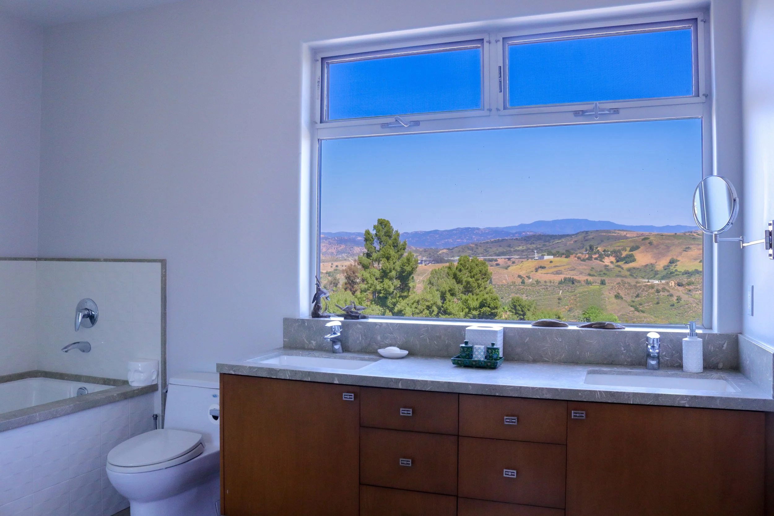 Bathroom with a large window showing a scenic landscape of trees and hills, a white toilet, and a vanity with a marble countertop, mirror, and various toiletries.