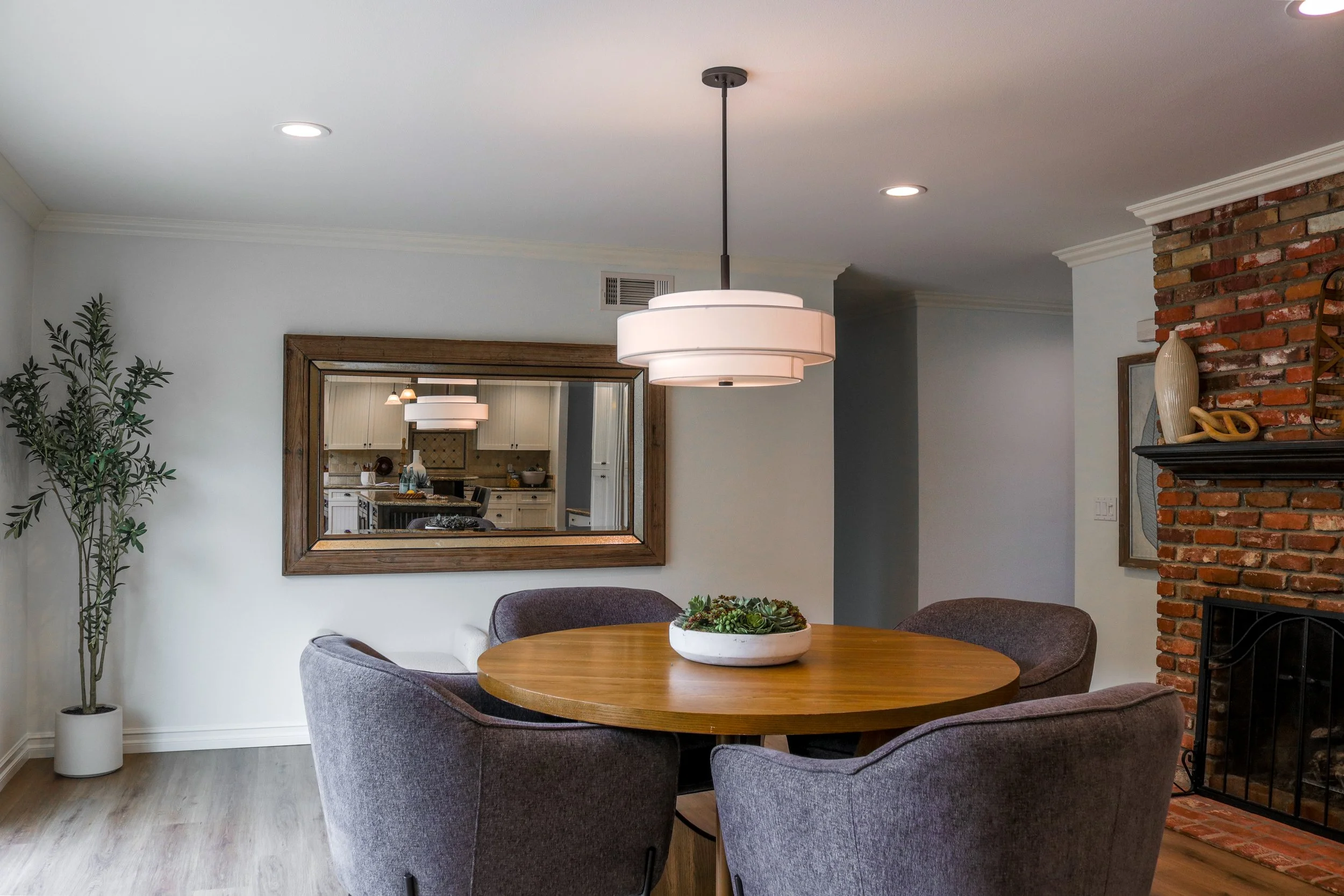 The image shows a cozy dining area with a round wooden table and four gray upholstered chairs. On the table, there is a white ceramic bowl with a green succulent plant. To the left, there is a tall potted plant with green leaves. In the background, a