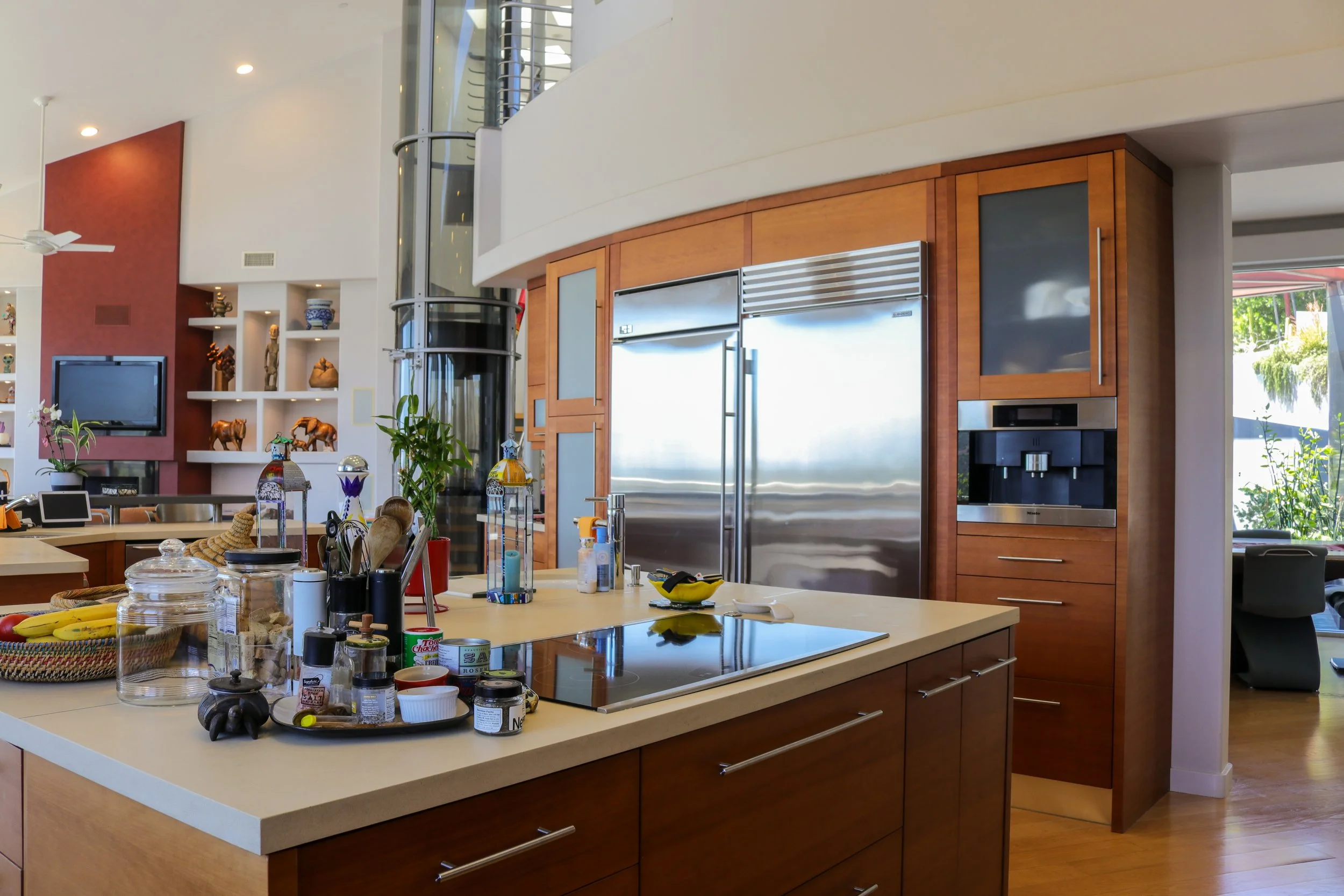 Modern kitchen with a large white island, wooden cabinets, stainless steel appliances, and decorative shelves. The counter has various kitchen items and a fruit bowl, with a TV mounted on the wall in the background.