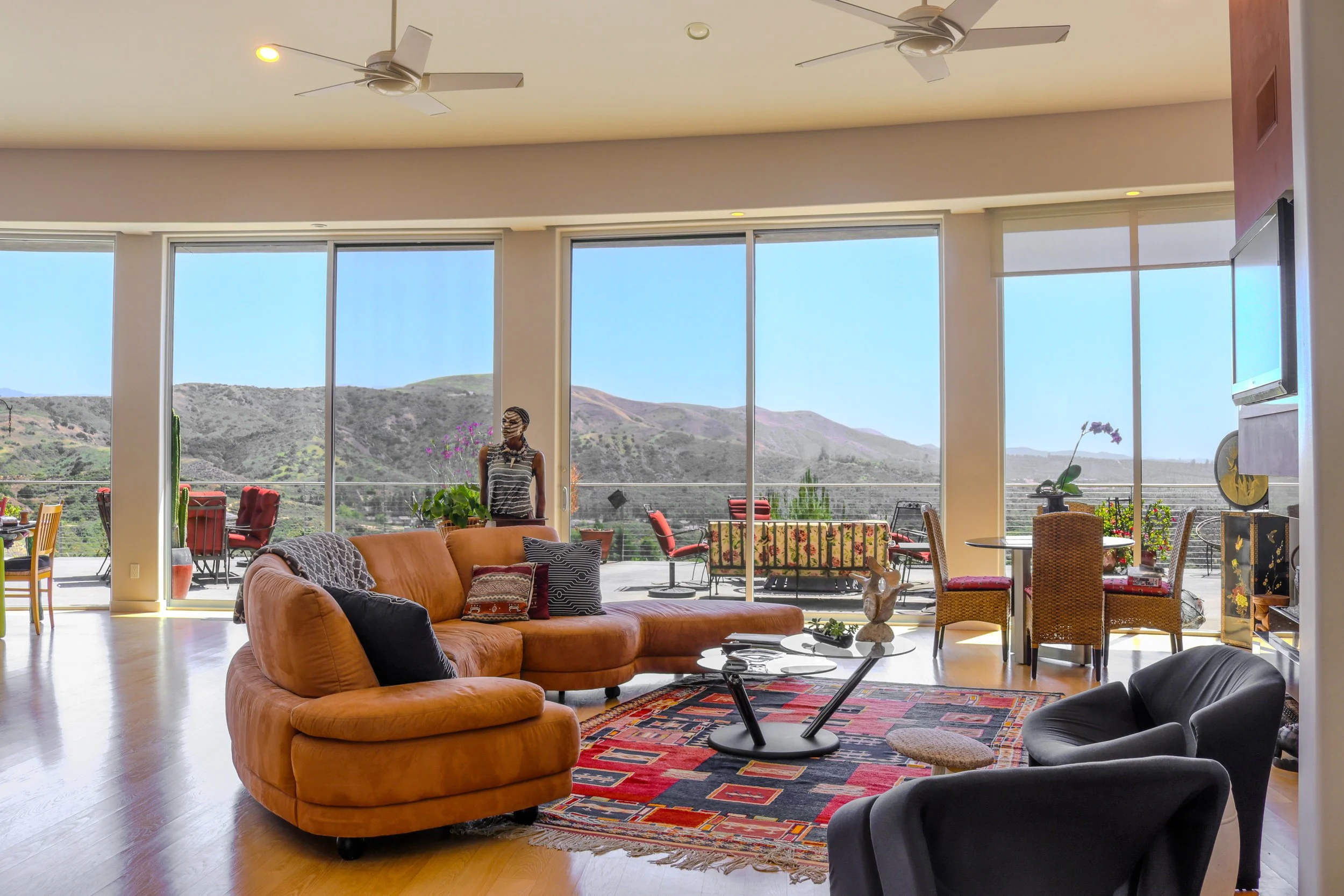 A spacious living room with large floor-to-ceiling windows showcasing a mountainous landscape. The room features a large orange sofa with throw pillows, a black armchair, a patterned area rug, and various side tables. The ceiling has two white ceiling fans, and there is a television mounted on a wall to the right.