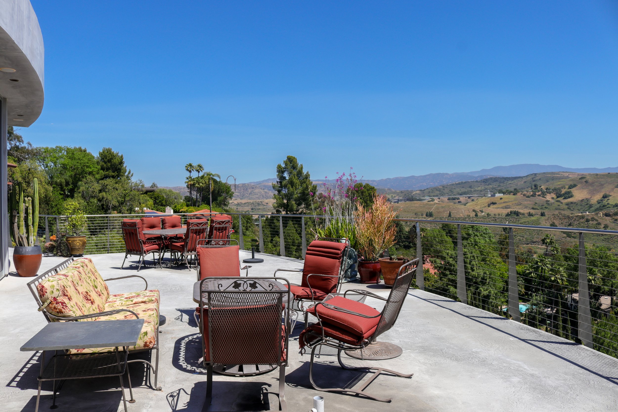 A spacious outdoor patio with colorful chairs and sofas, potted plants, and a view of rolling hills and mountains under a clear blue sky.