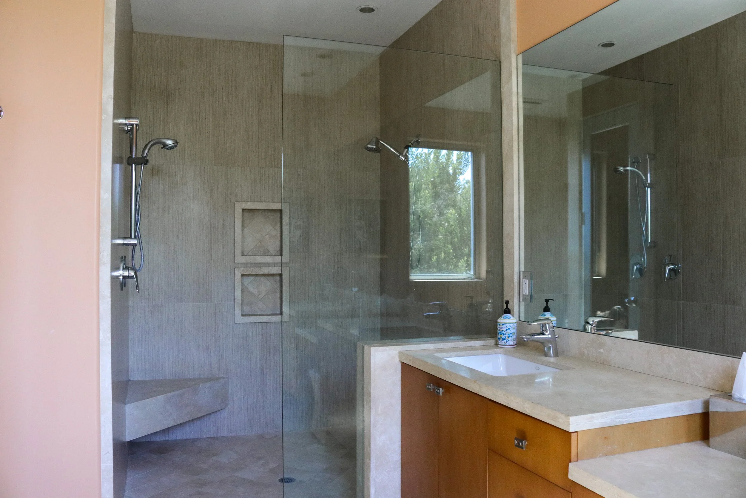 Modern bathroom with a glass-enclosed shower area featuring two shower heads, a small window with a view of trees, a mirror above the sink, two dispensers, and a vanity with wooden cabinets.