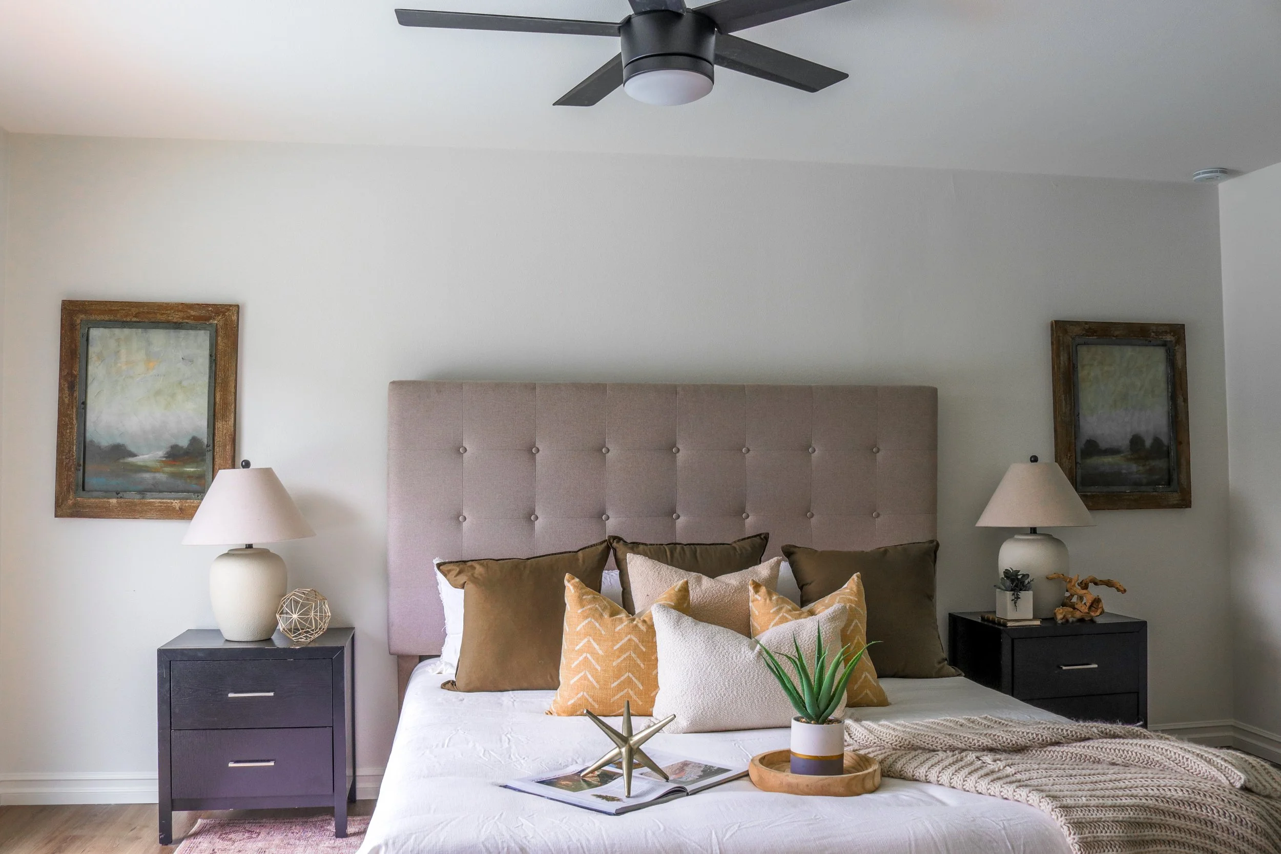 A bedroom with a beige upholstered headboard, two side tables with white lamps, decorative pillows on the bed, artwork on the walls, a ceiling fan, a potted plant, and a tray with decor on the bed.