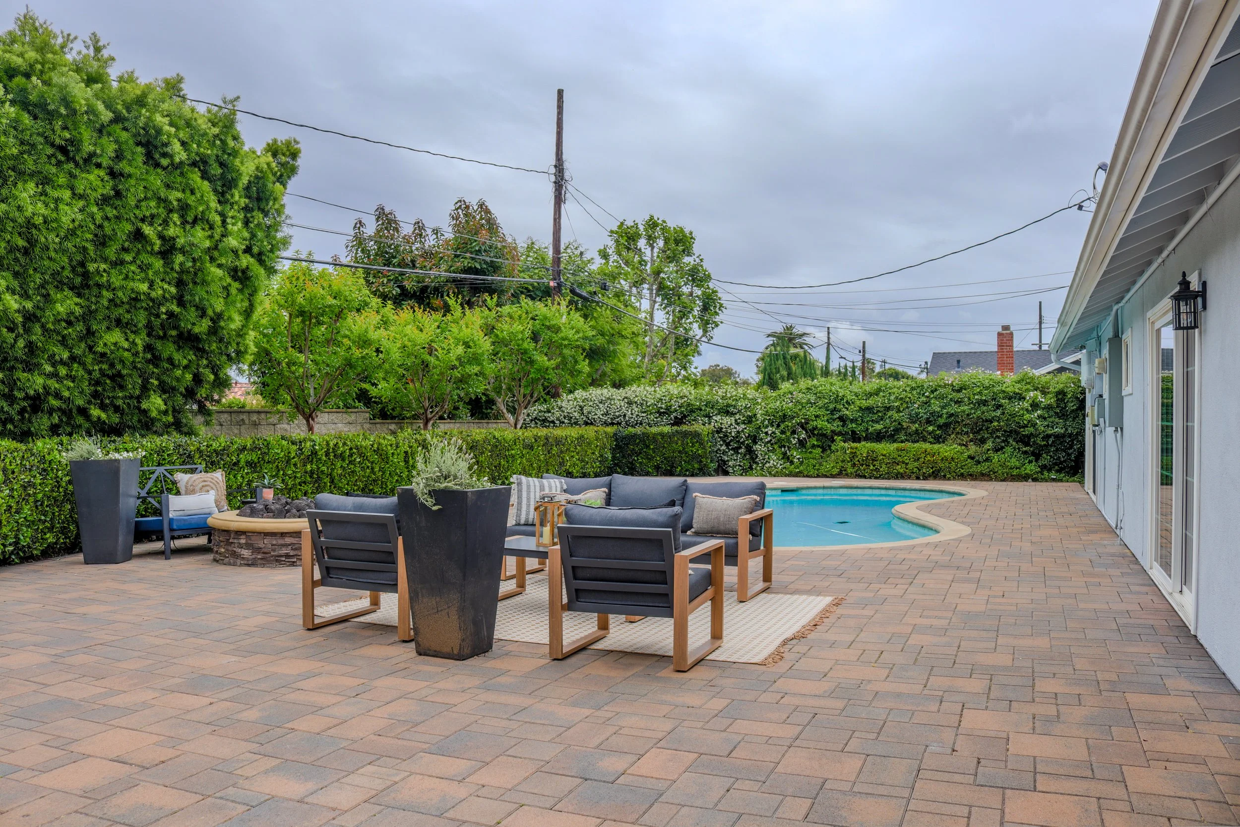 A backyard with brick patio, outdoor seating, potted plants, swimming pool, surrounded by green trees and bushes, overcast sky.