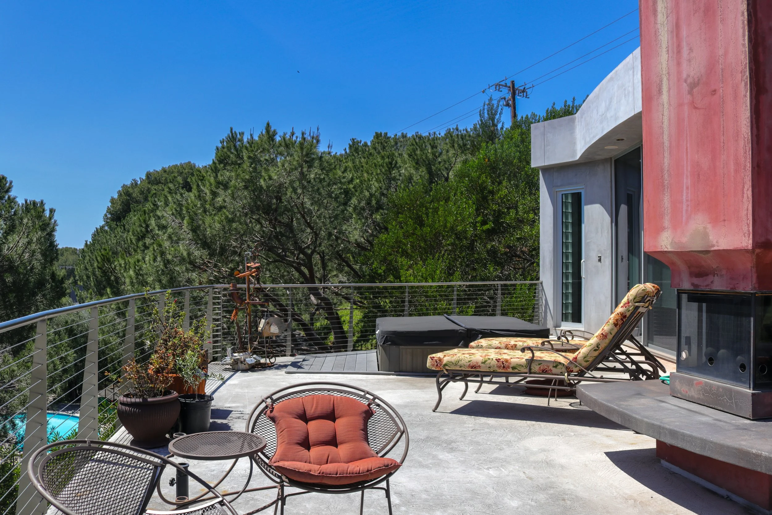 An outdoor rooftop patio with a concrete floor, lounge seating with floral cushions, potted plants, and a view of trees, with a modern building wall on one side and a railing on the edge.