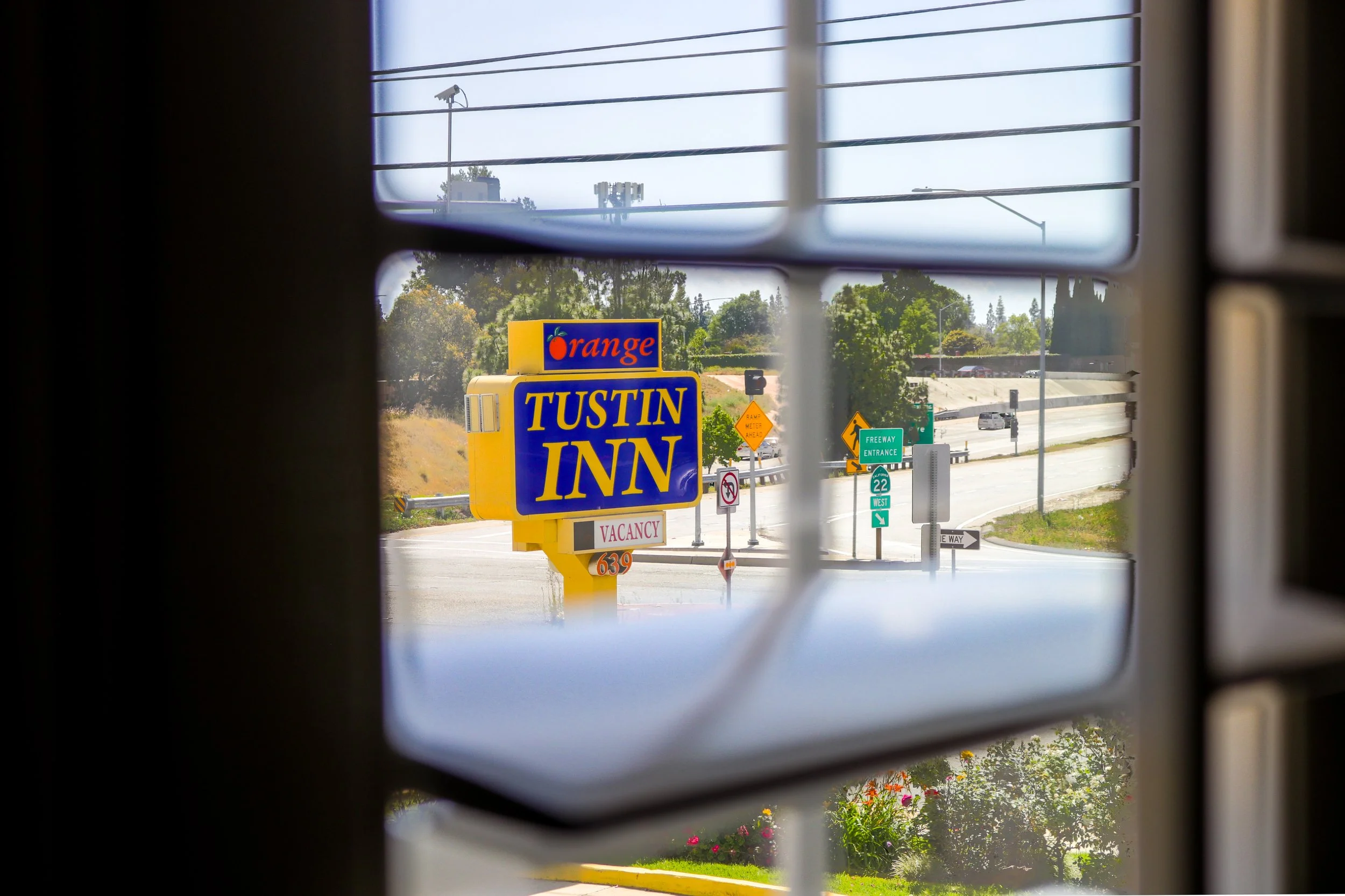 View of a roadside sign for Tustin Inn with purple and yellow colors, partially seen through a window with blinds, showing a street with traffic signs and trees in the background.