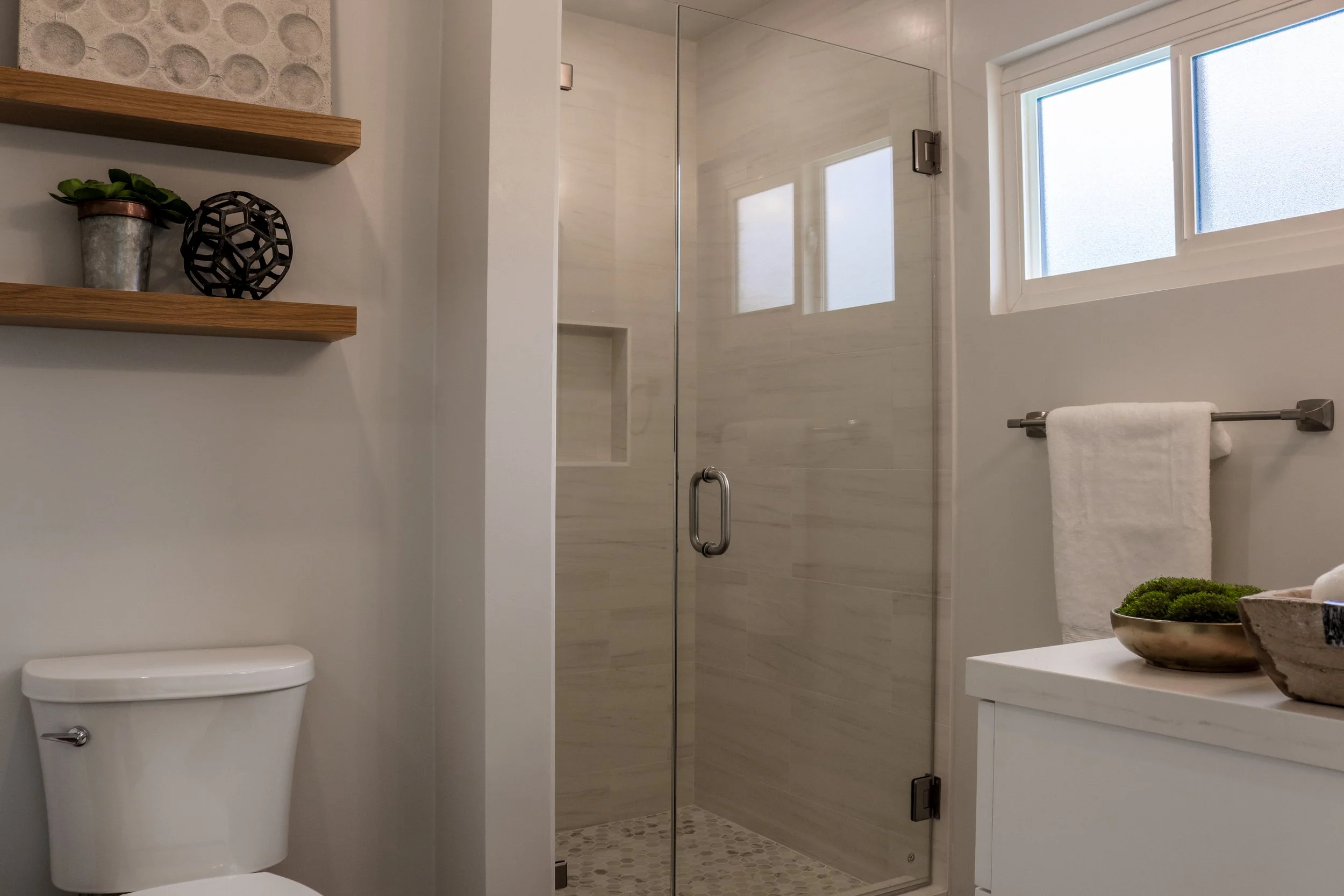 A modern bathroom with a glass shower door, a toilet, a window, a towel rack with a white towel, and wooden shelves with decorative items including a plant and a black geometric sculpture.