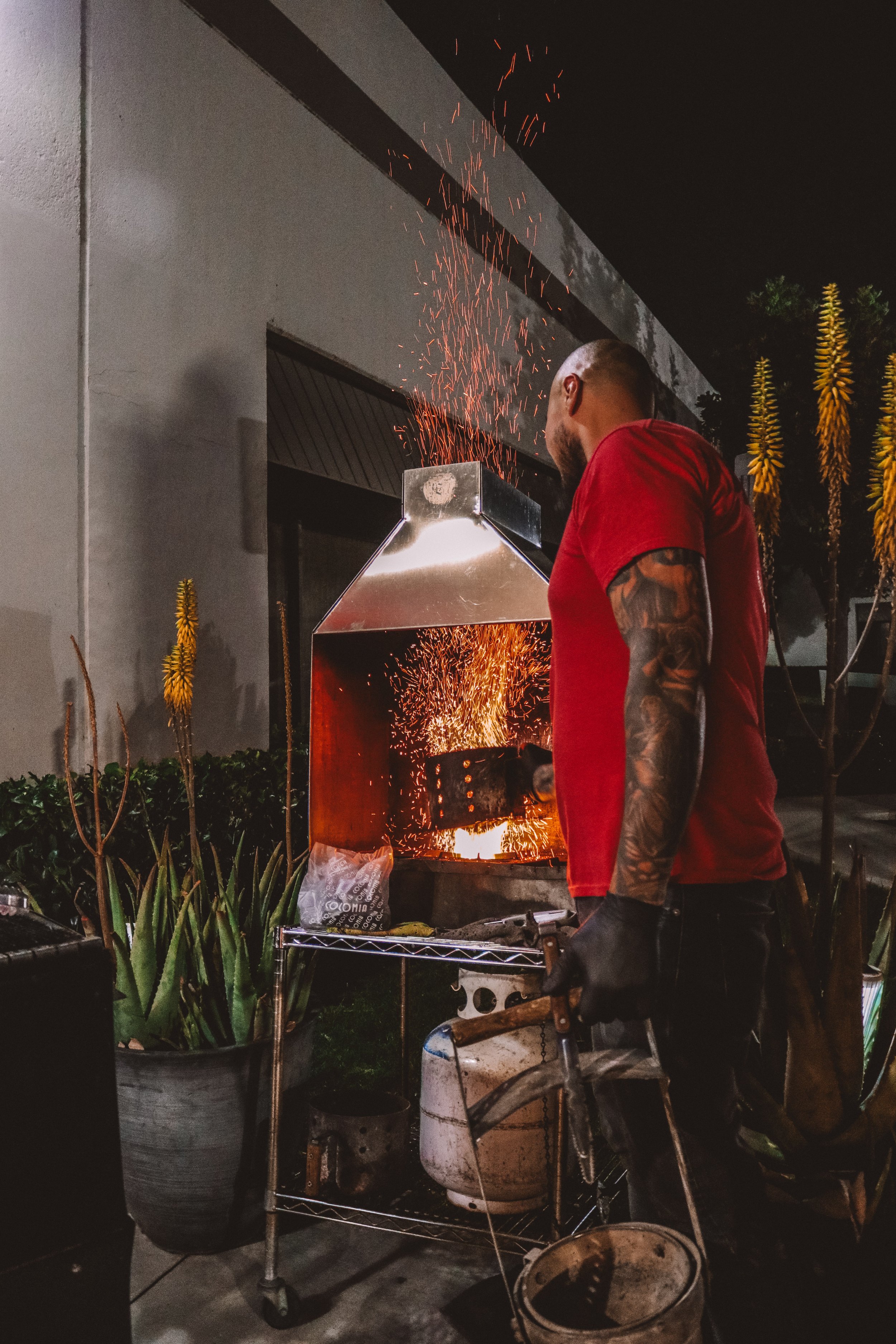 A man wearing a red shirt and black gloves is using a torch to melt or cut metal outside at night. Sparks are flying from the metal, and he appears focused on his work. There are plants and potted greenery nearby, and a metal cart with supplies and t