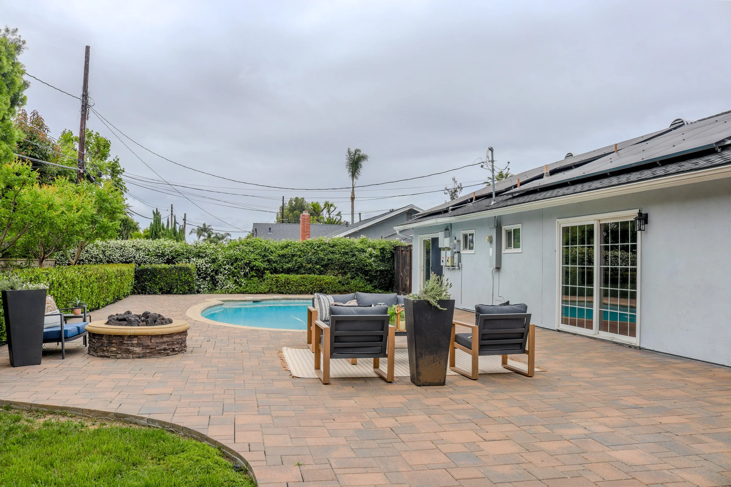 Backyard patio with seating area, small pool, fire pit, surrounded by green bushes and trees, with a house featuring sliding glass doors and gray roof, overcast sky.