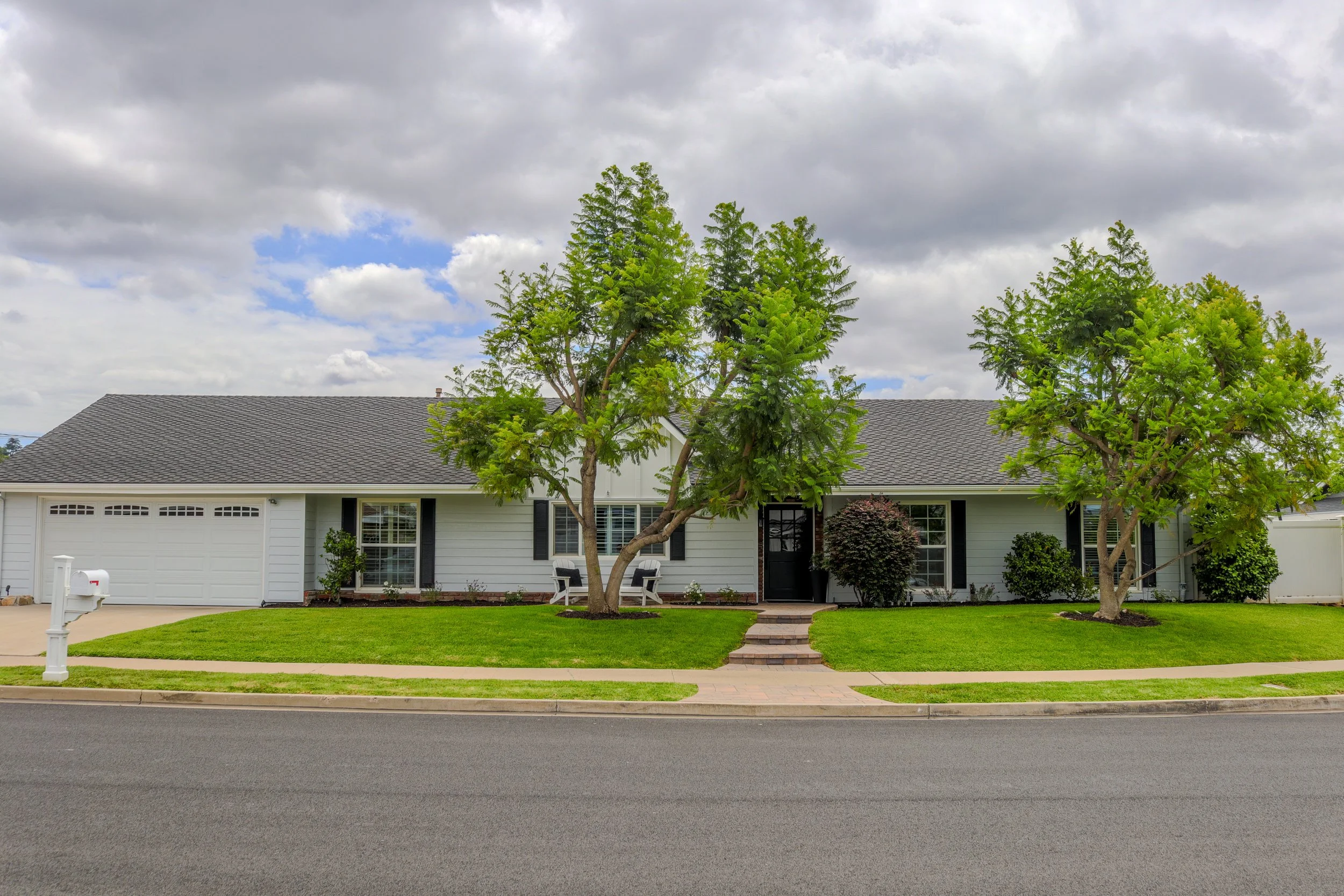 Front view of a suburban house with a gray shingle roof, white exterior walls, and black window shutters. In front of the house, there are three green trees, a well-maintained lawn, and a concrete pathway leading to the front door. A white mailbox is
