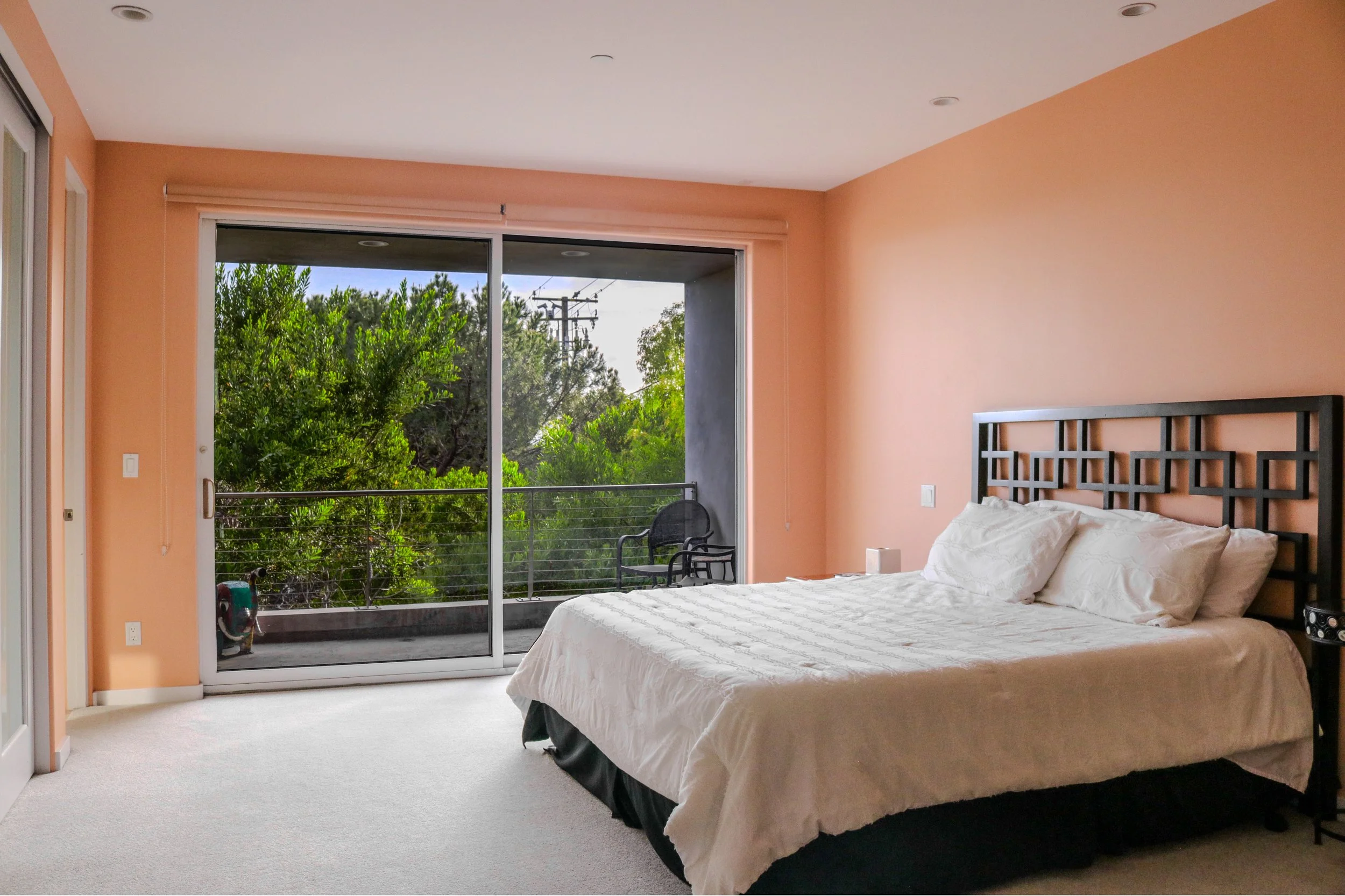 A bedroom with peach-colored walls, a large window with sliding glass doors leading to a balcony, and a bed with white bedding. There is outdoor greenery visible through the window, and a chair on the balcony.