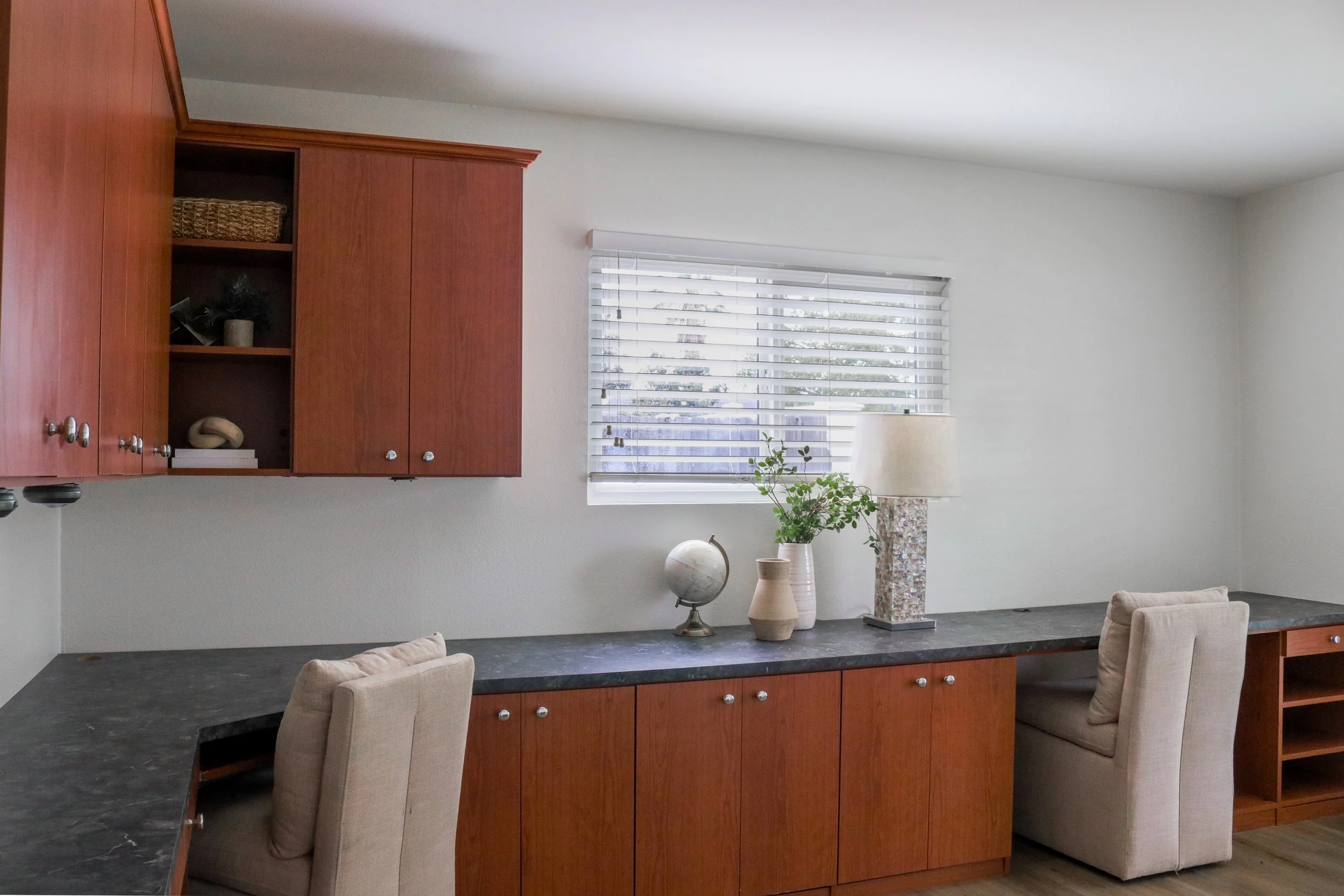 Living room with a large wooden cabinet, marble countertop, potted plants, a globe, a lamp, and beige chairs near a window with white blinds.