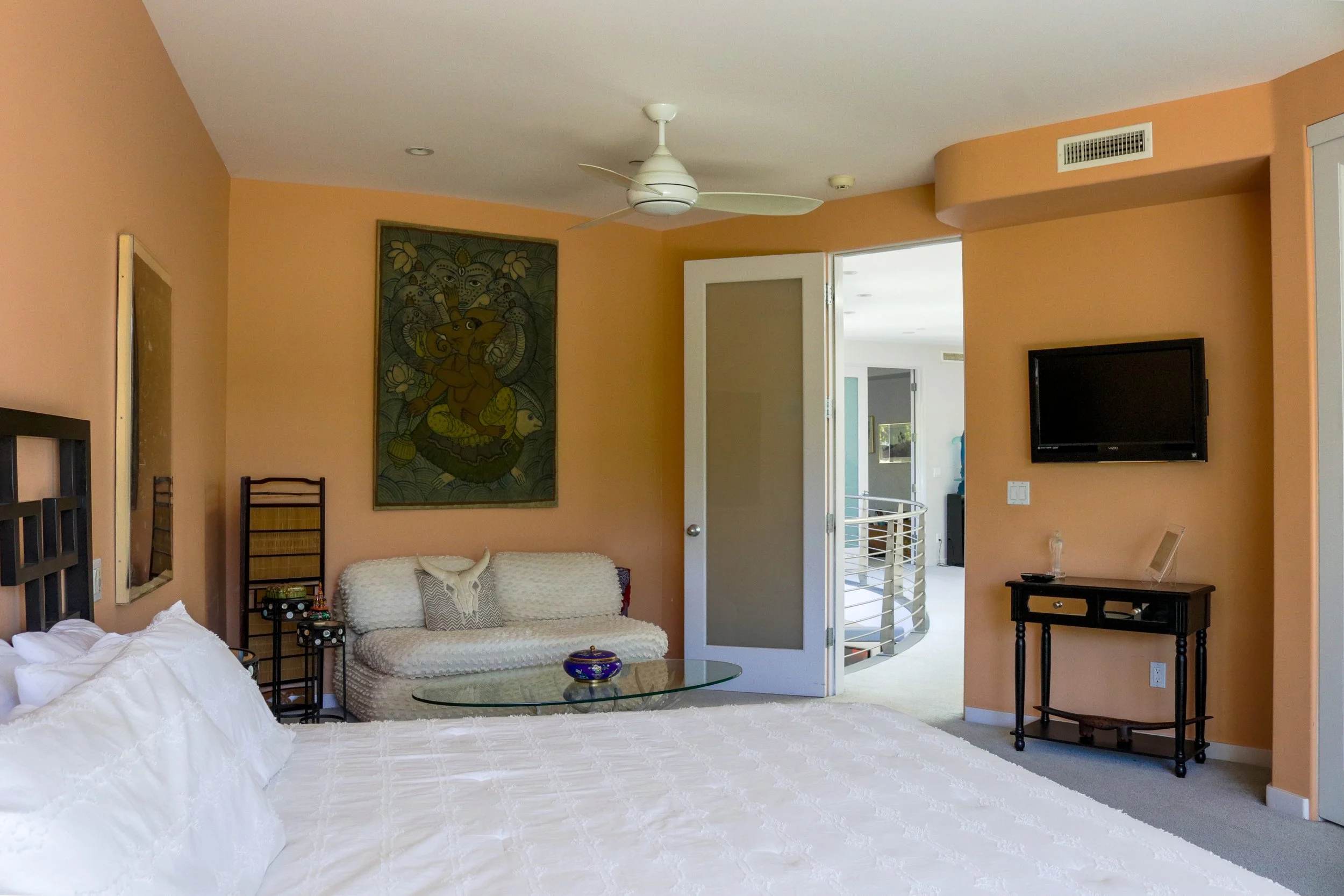 Living room with peach-colored walls, a ceiling fan, a flat-screen TV, artwork on the wall, a white sofa with a blanket, a glass table, and a door leading to a balcony or patio.
