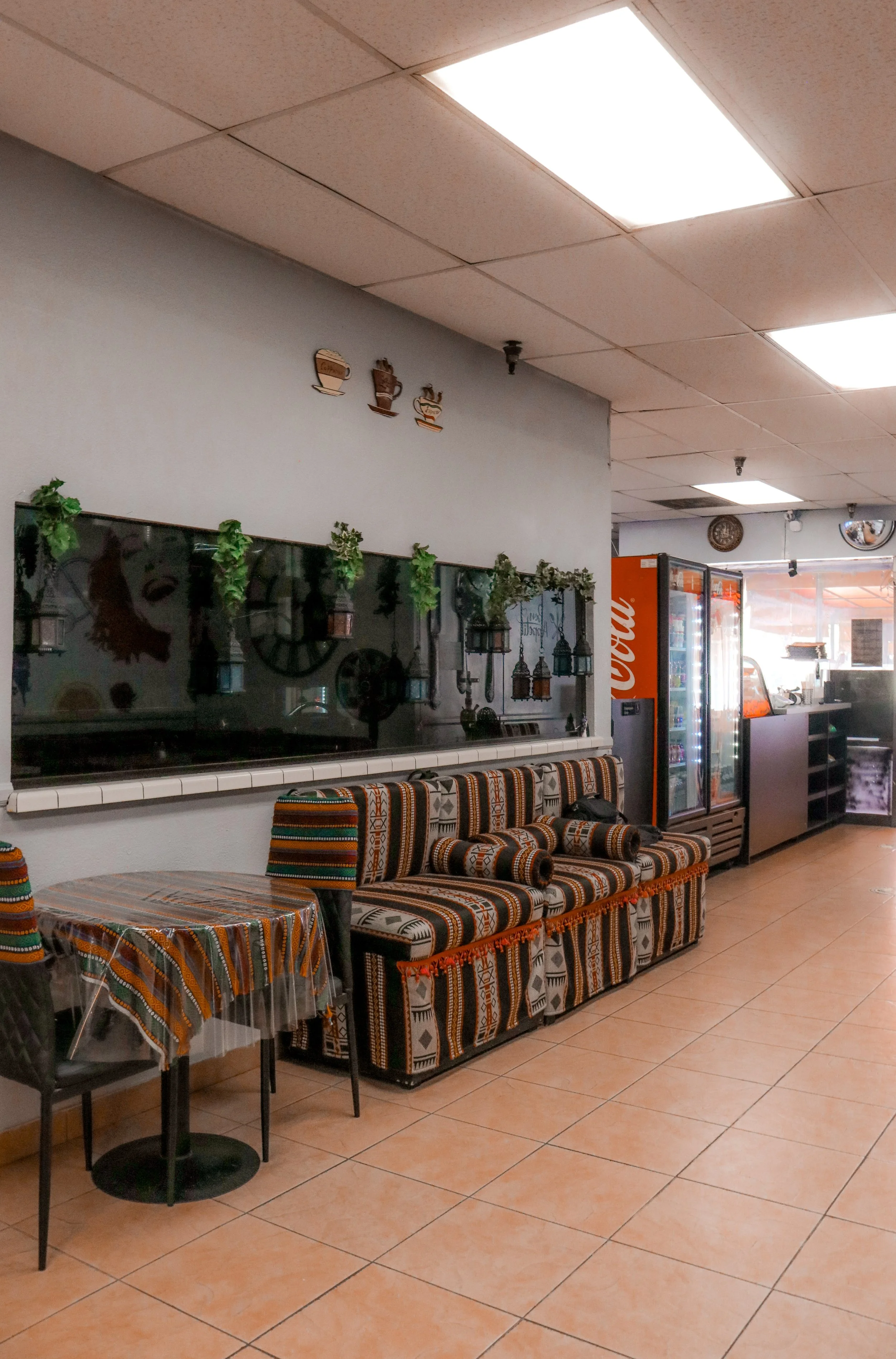 Interior of a cafe or restaurant with patterned cushioned seating, a round table with a patterned cloth, green decorative plants on a mirror wall, and a vending machine in the background.
