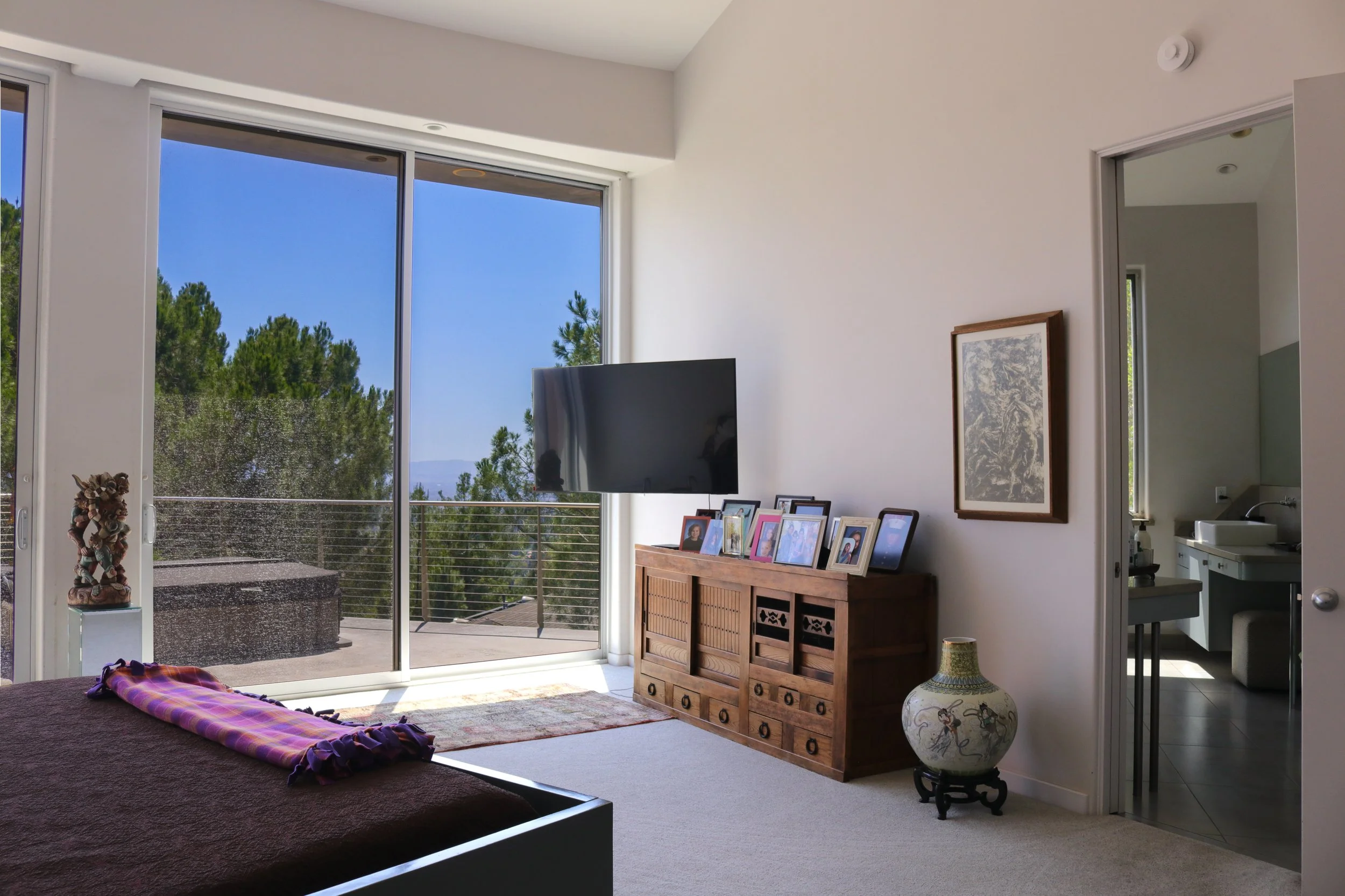 Living room with large sliding glass door showing balcony and trees outside, a wooden cabinet with photo frames, a mounted TV, and a table with decorative items.