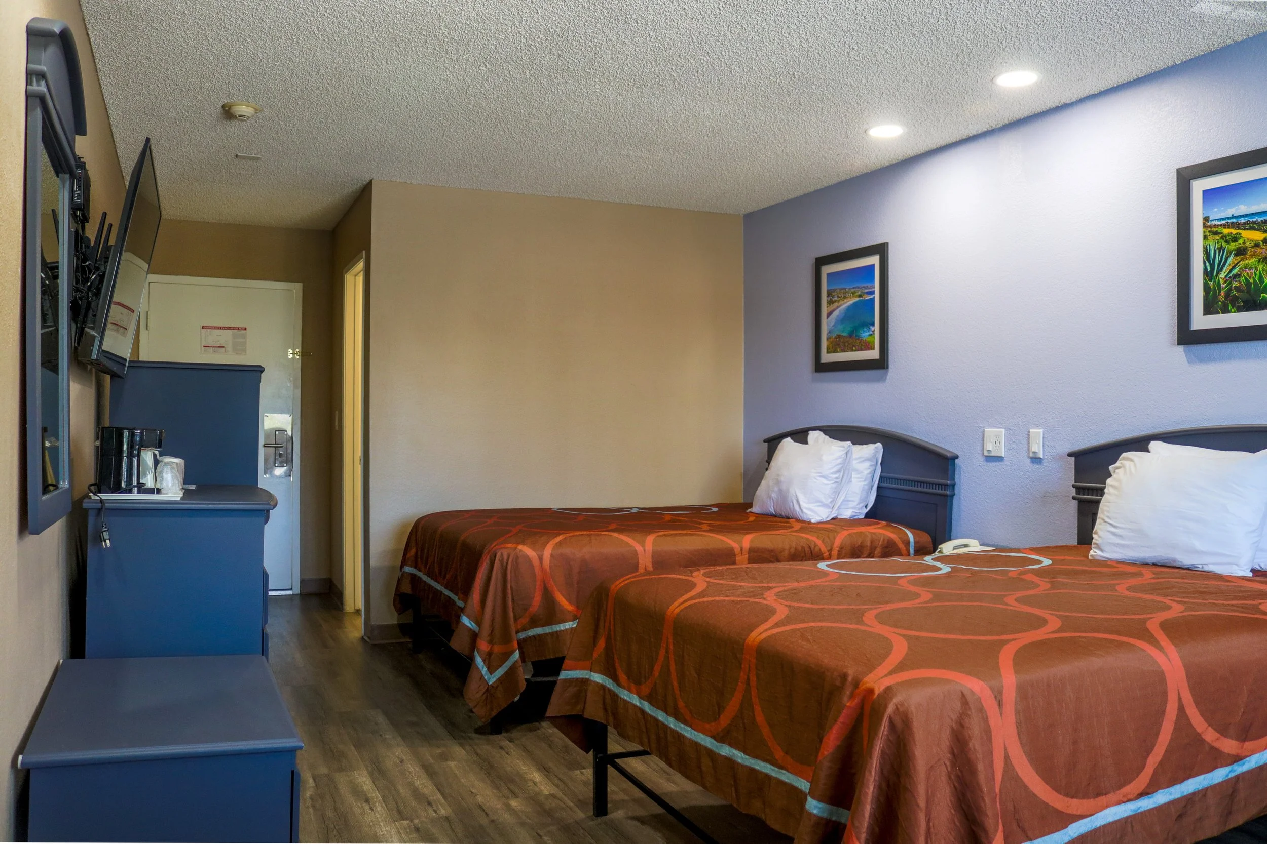 Two beds with orange and brown patterned bedspreads and white pillows in a hotel room. The room has framed landscape photographs on the wall, a blue dresser, and a wall-mounted flat-screen TV.