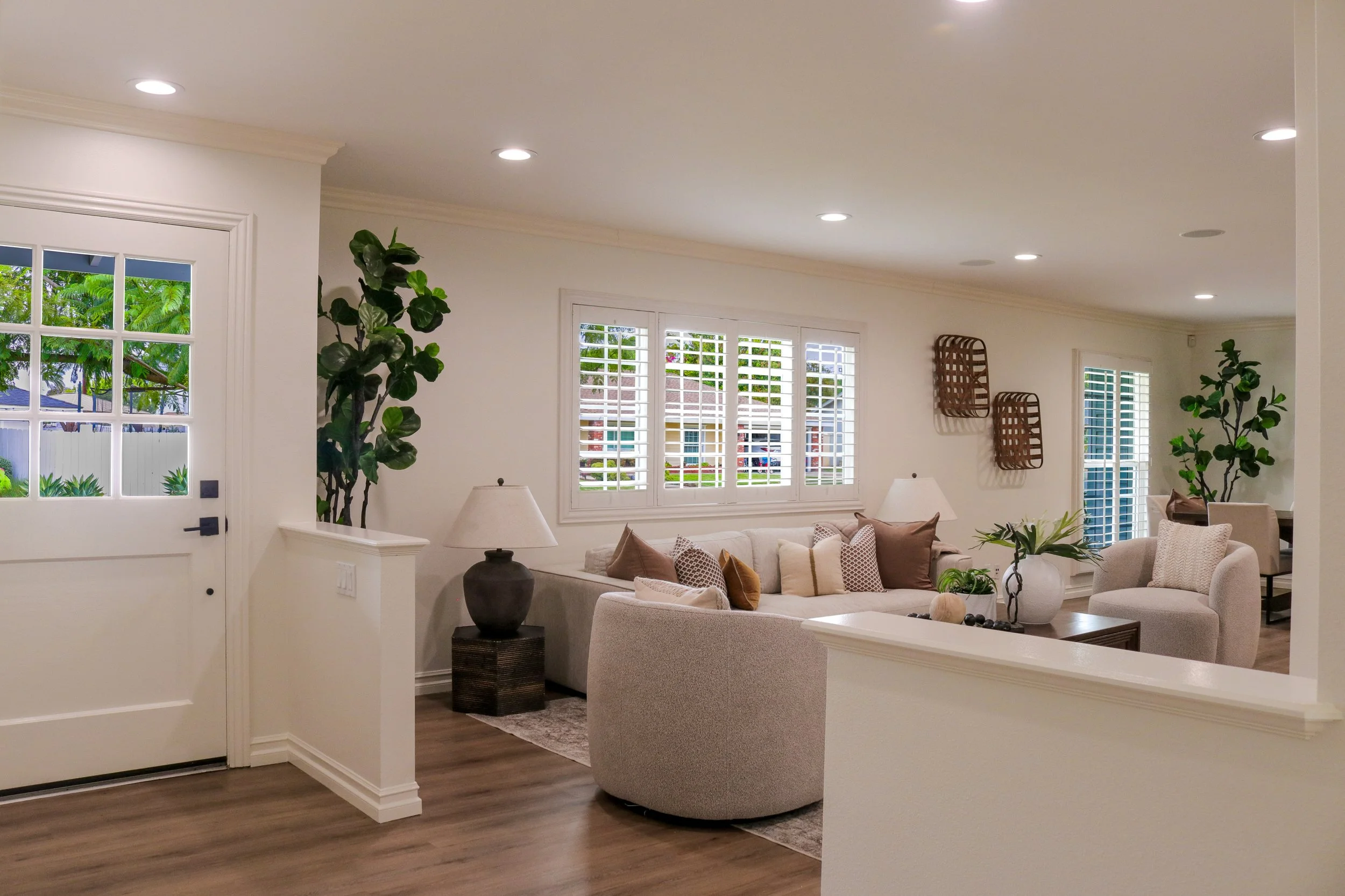 A cozy living room with a white sofa decorated with beige and patterned throw pillows, a dark wood side table with a large dark vase, and a lamp. Large windows with white shutters allow natural light, and potted plants add greenery. The ceiling has r