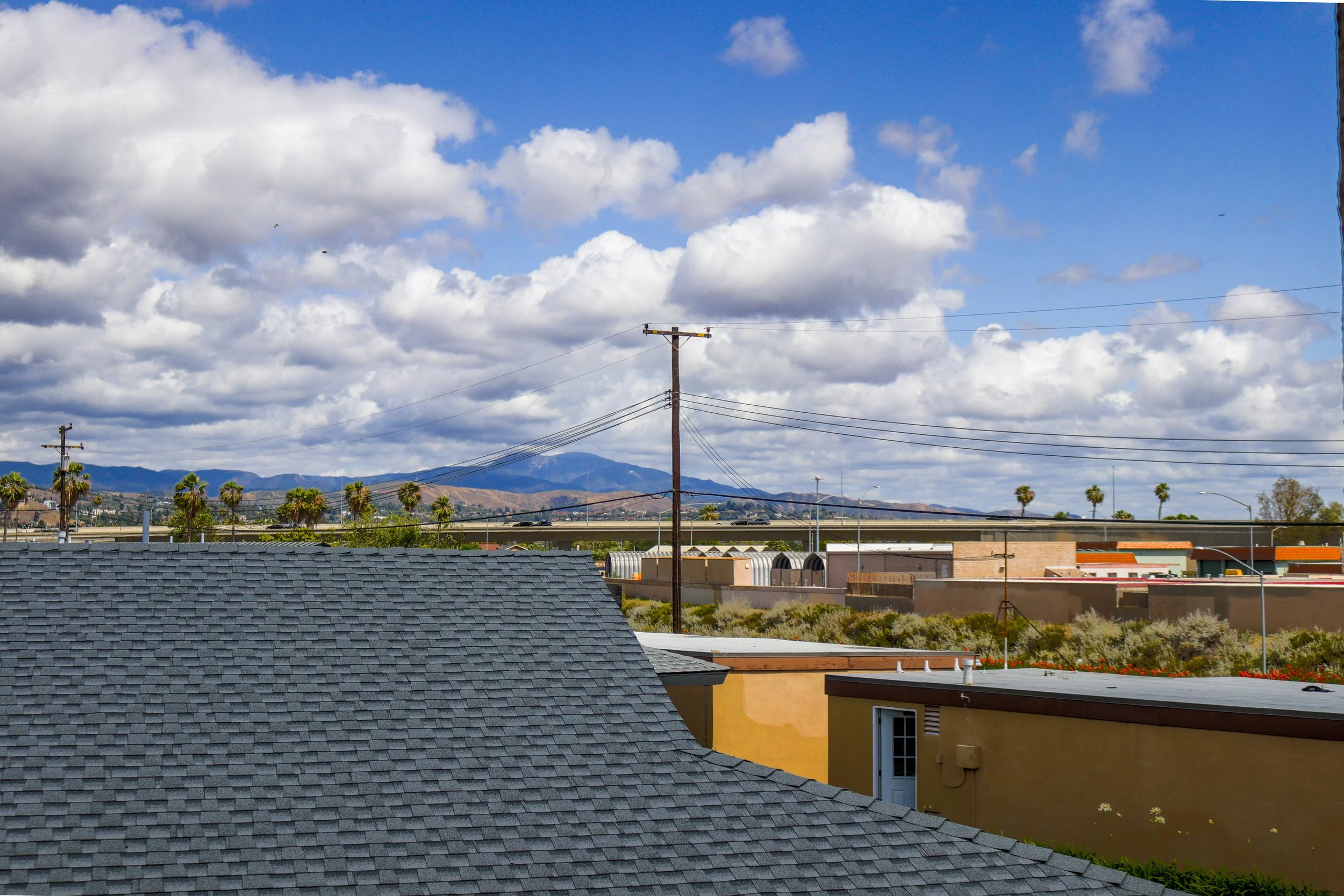View of suburban neighborhood with rooftops, power lines, palm trees, and mountains in the distance under a partly cloudy sky.
