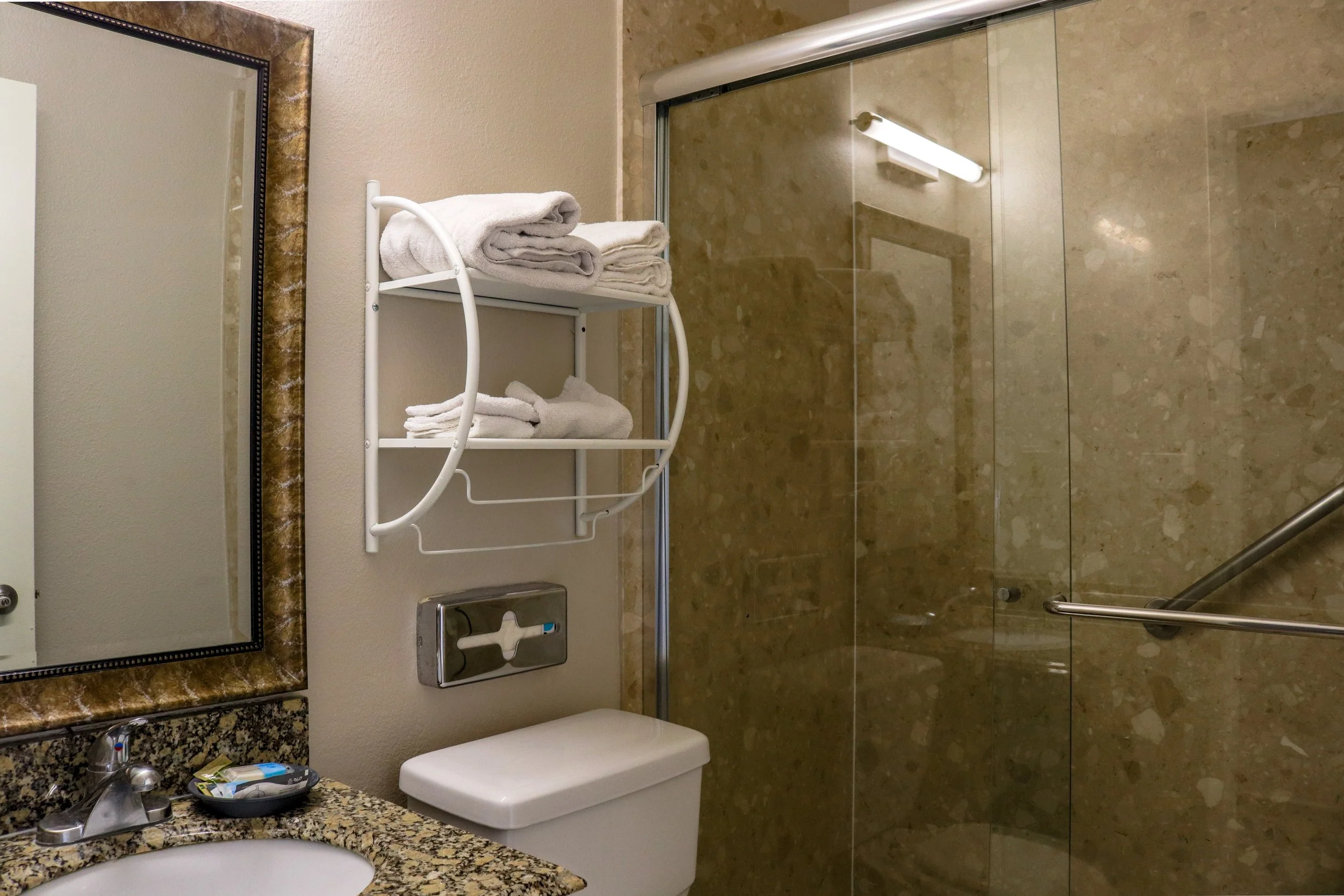 A bathroom with a granite countertop, a mirror with a decorative frame, a small sink, and a toilet, alongside a shower with a glass door and a towel rack with rolled-up towels.