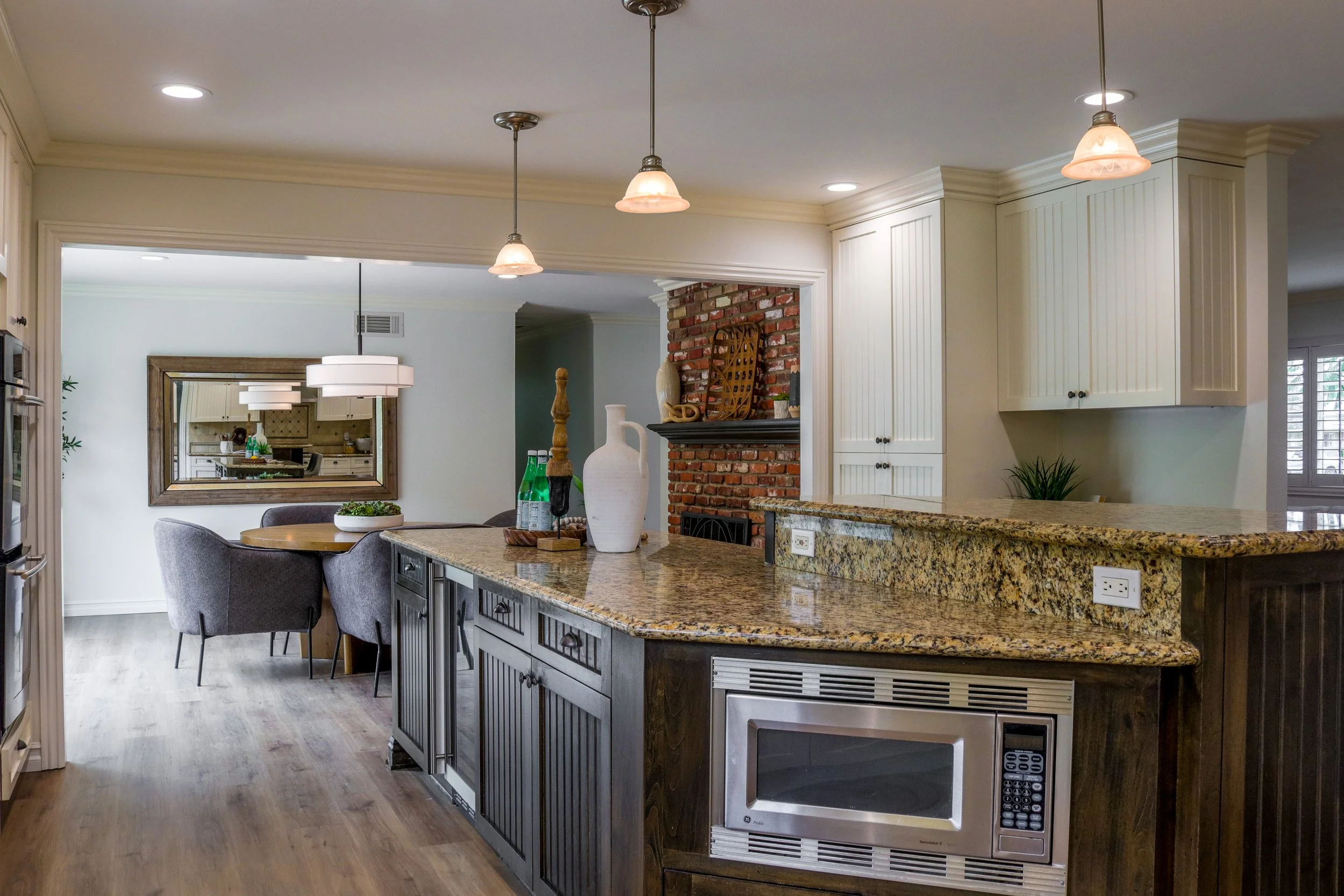 A modern kitchen with a large granite island, white cabinets, a brick fireplace, and a dining area with a round table and gray chairs.