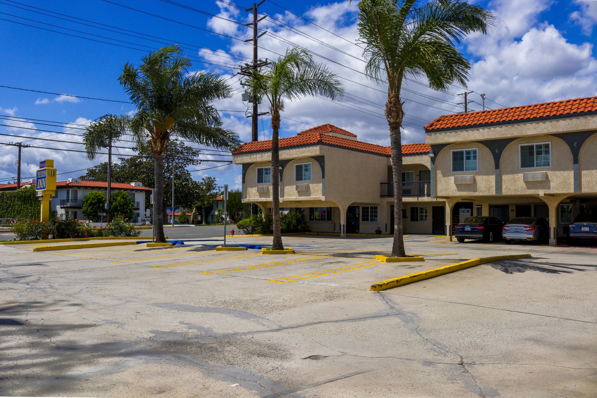 Empty parking lot in front of beige apartment buildings with red tile roofs, tall palm trees, and power lines against a partly cloudy blue sky.