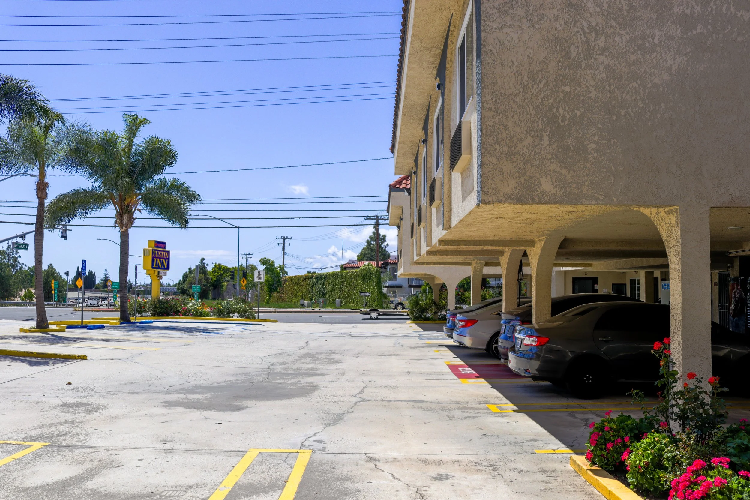 Empty parking lot outside a beige Spanish-style building with palm trees and a sign for Tustin Inn. Clear blue sky and power lines overhead.