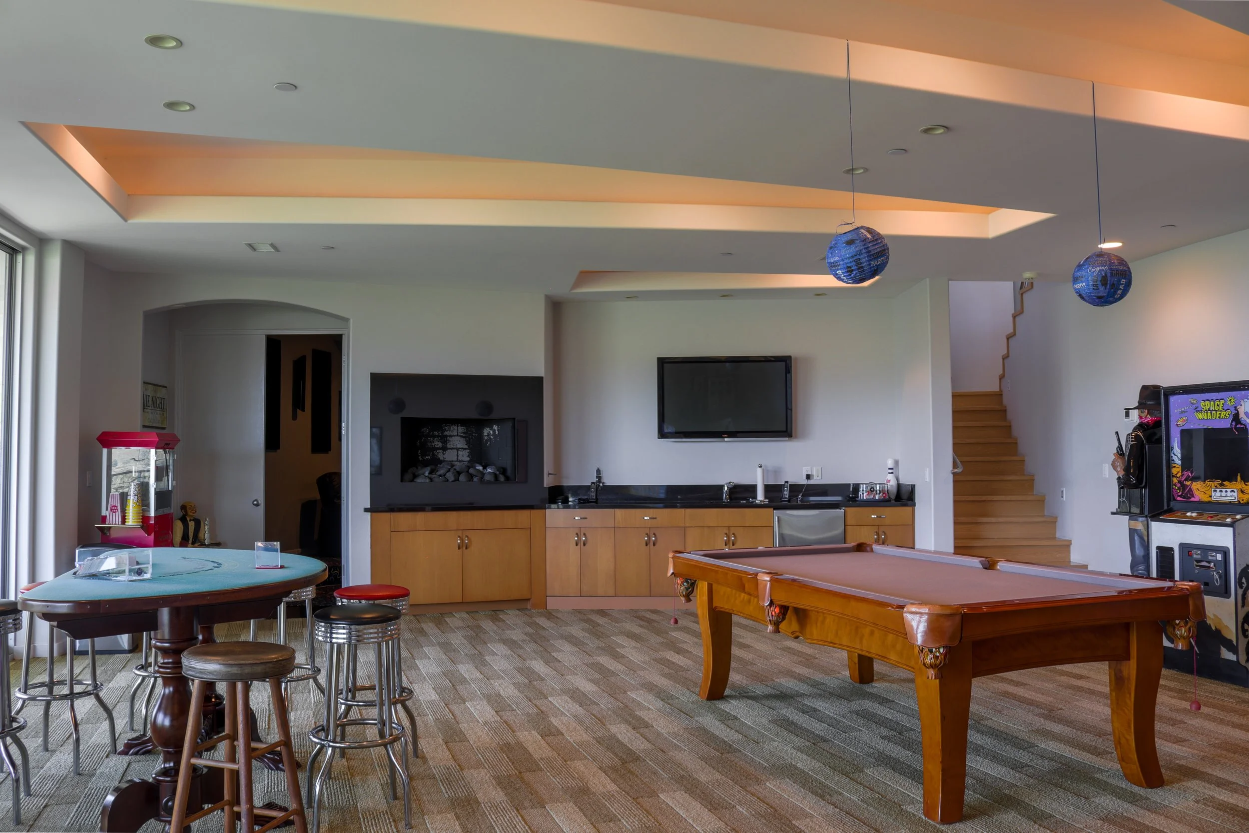 A spacious game room with a pool table, a dartboard, and various furniture. The ceiling has recessed lighting and decorative lighting features. The room is illuminated by natural light from large windows on the left side.