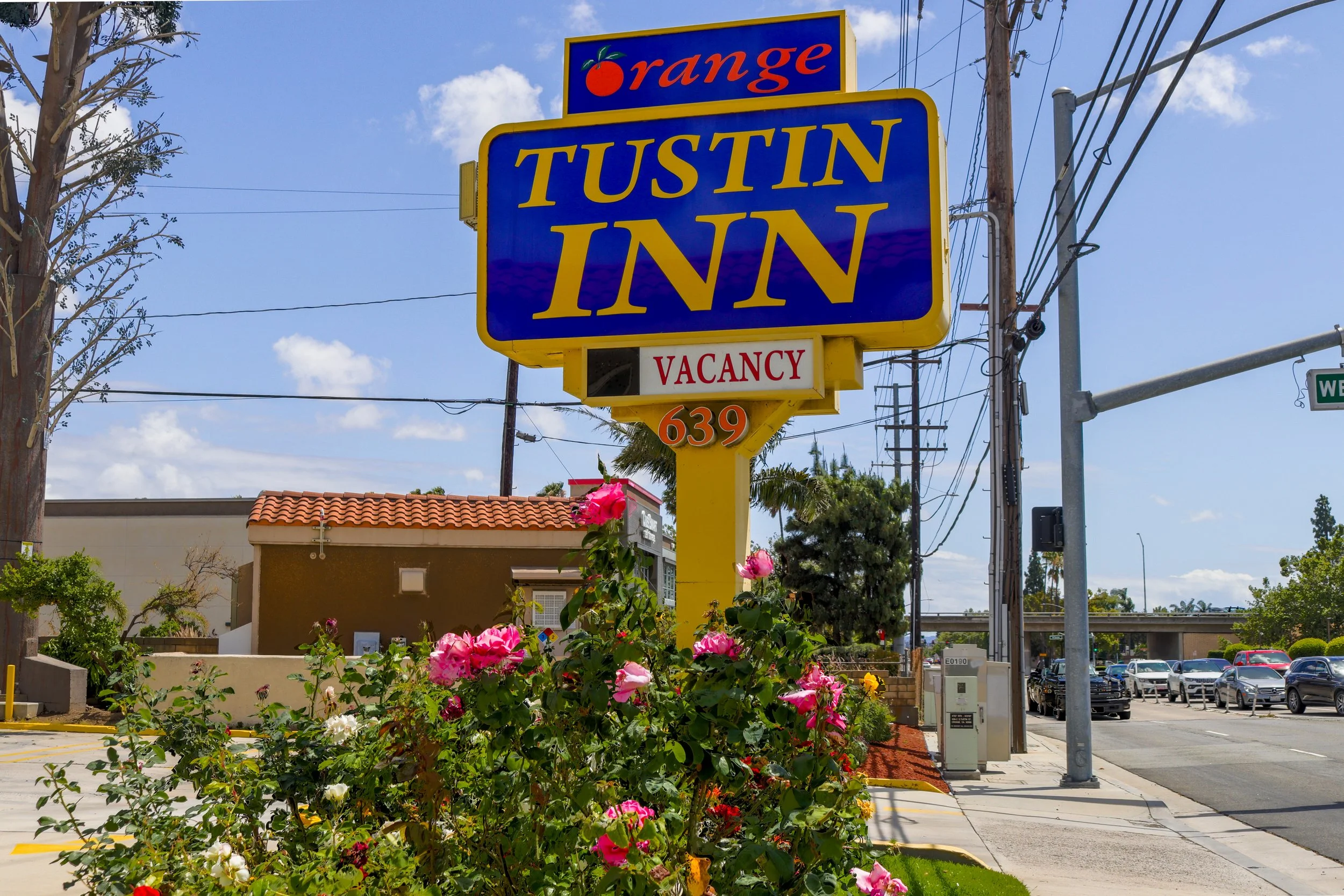 Blue and yellow sign for Tustin Inn with a smaller sign below indicating vacancy, located in a commercial area with flowers in the foreground and trees, power lines, and a parking lot in the background.