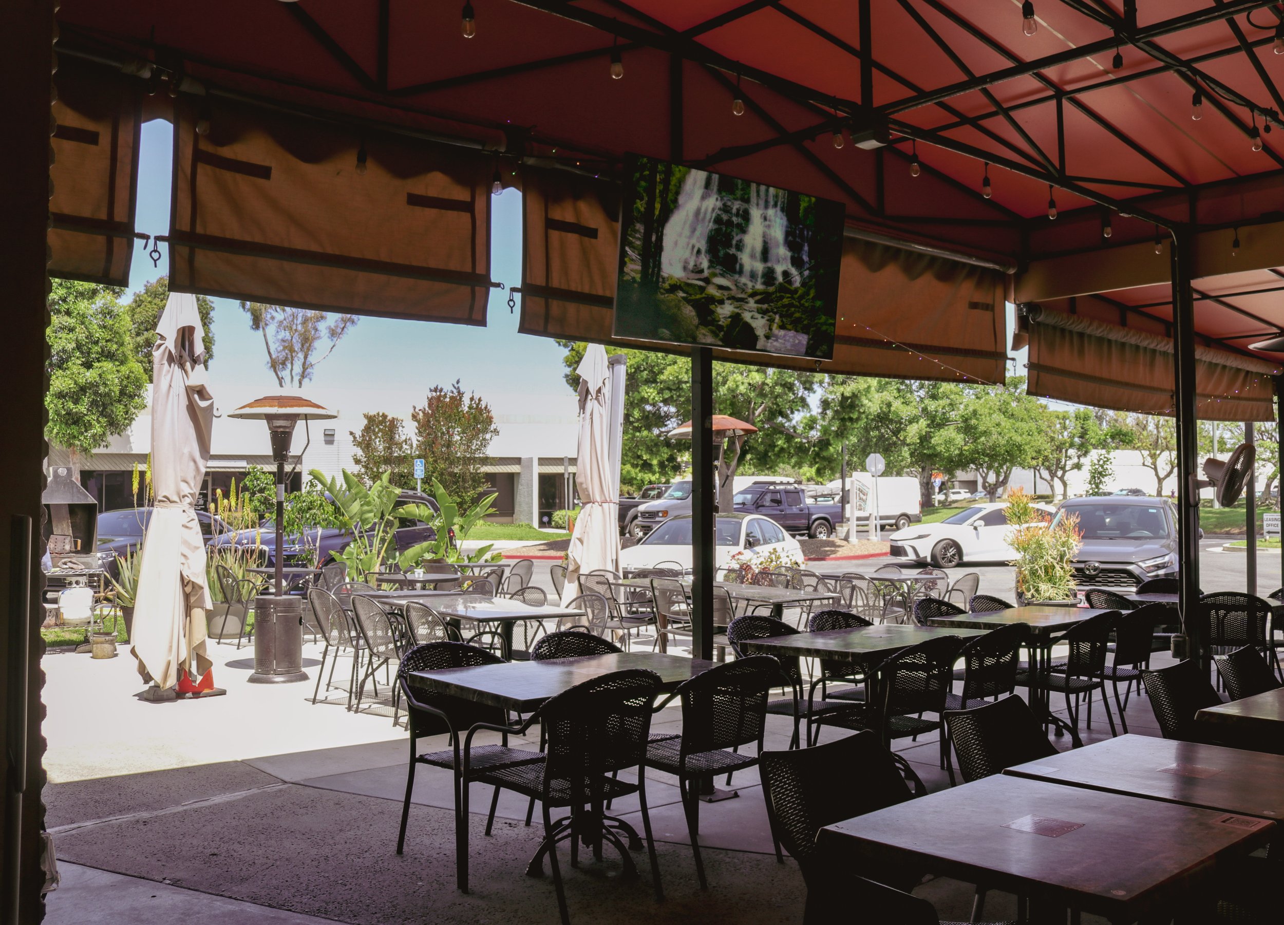 View of an outdoor patio dining area with black chairs and tables, part of a restaurant, with umbrellas, trees, and parked cars visible in the background, and a large water fountain on a screen inside.