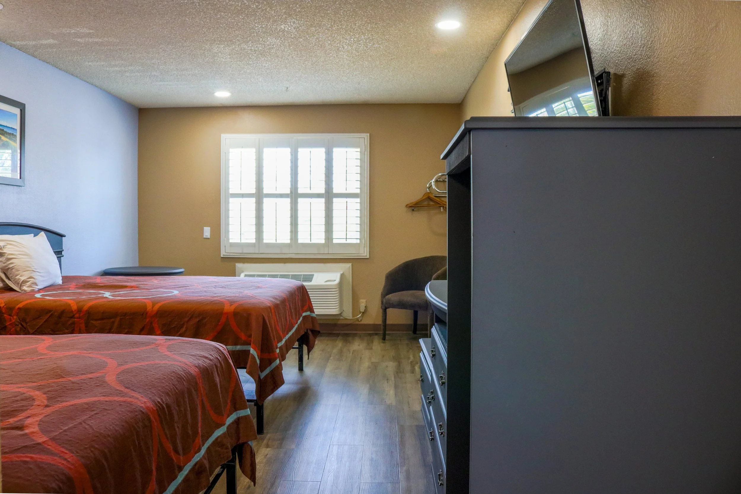 A hotel room with two beds, one with an orange and red bedspread, a window with shutters letting in natural light, a wall-mounted TV on a gray cabinet, and a small ergonomic chair.