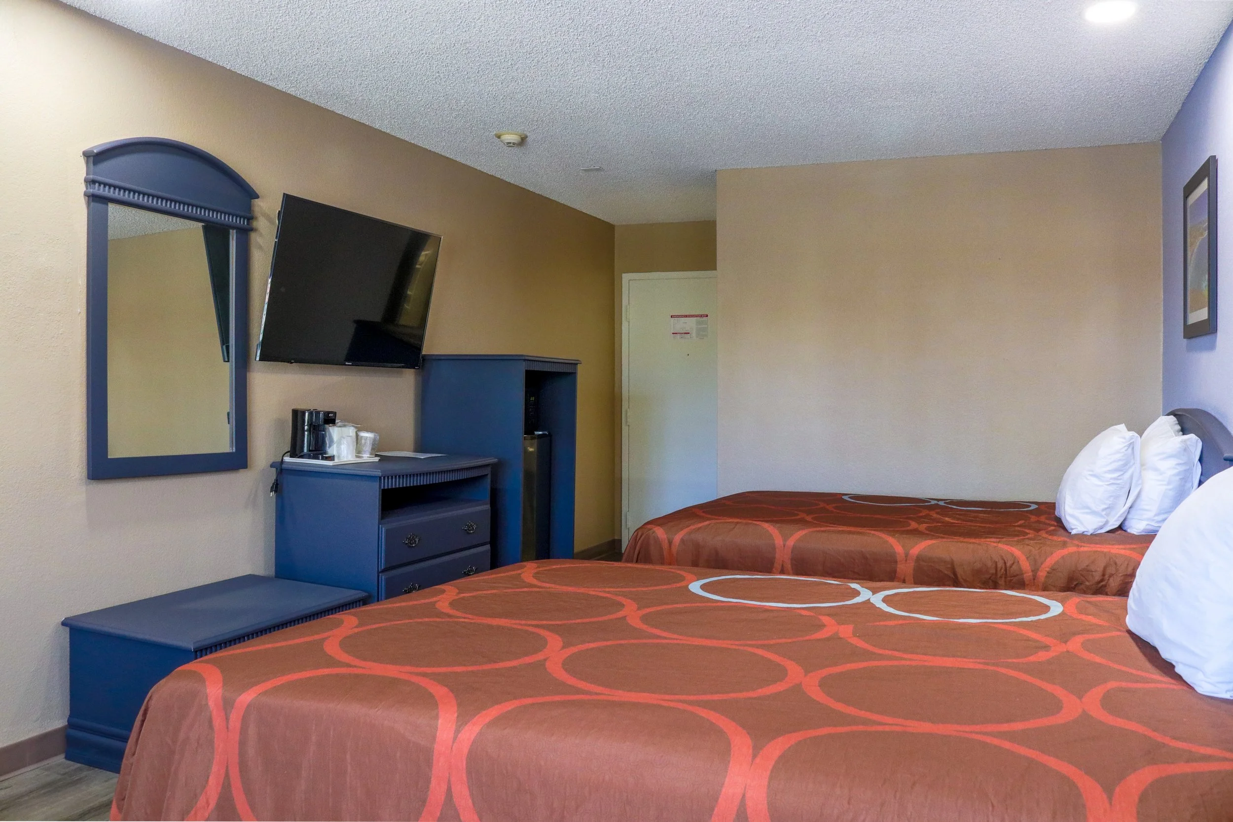 Two hotel beds with orange patterned bedspreads and white pillows in a room with beige and blue walls, a wall-mounted TV, a mirror, and a breakfast station.