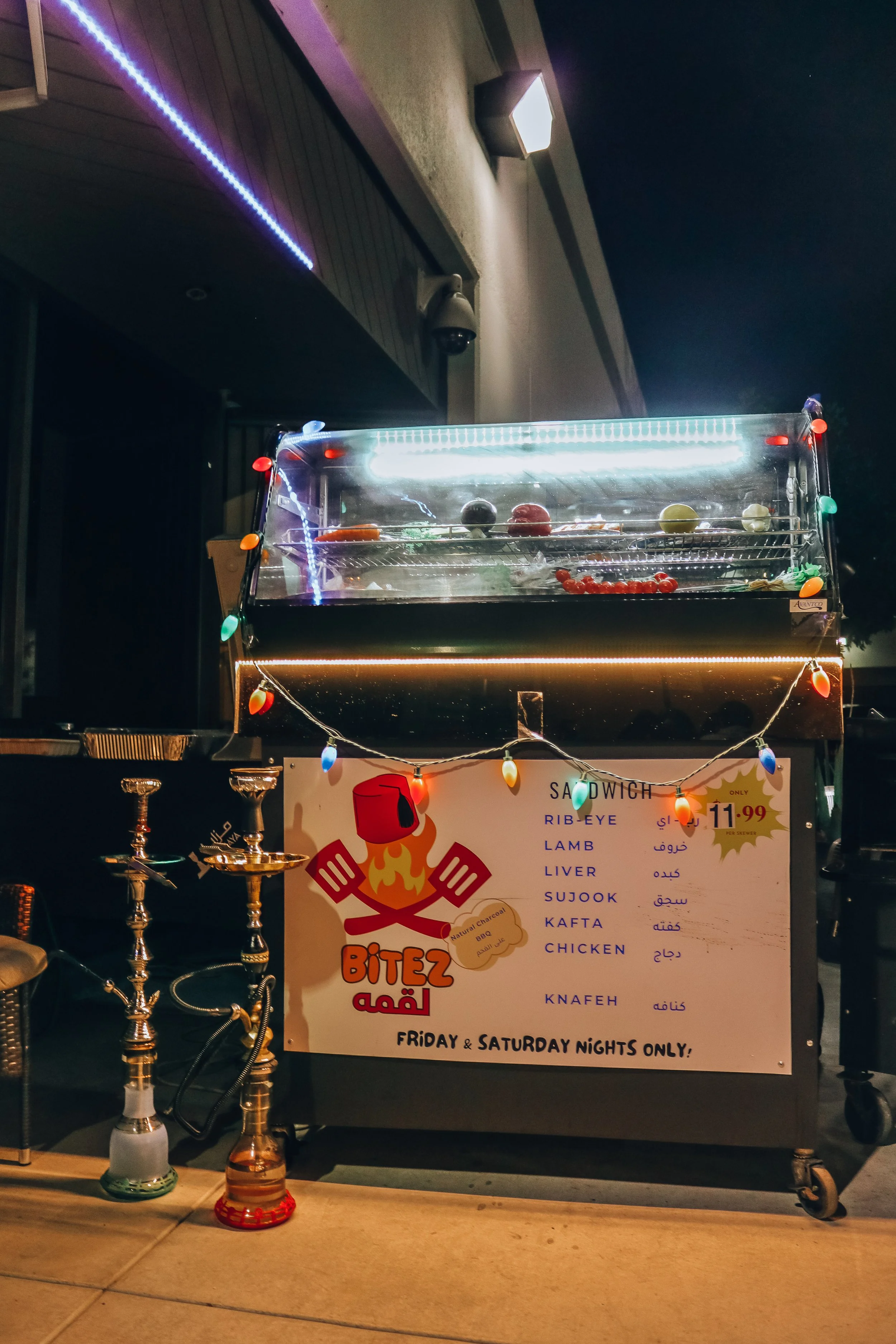 A food cart with a sign advertising different types of kebabs including rib-eye, lamb, liver, sujook, kafta, chicken, and knafeh, with the text that it is only open on Friday and Saturday nights. The cart is decorated with colorful string lights and 