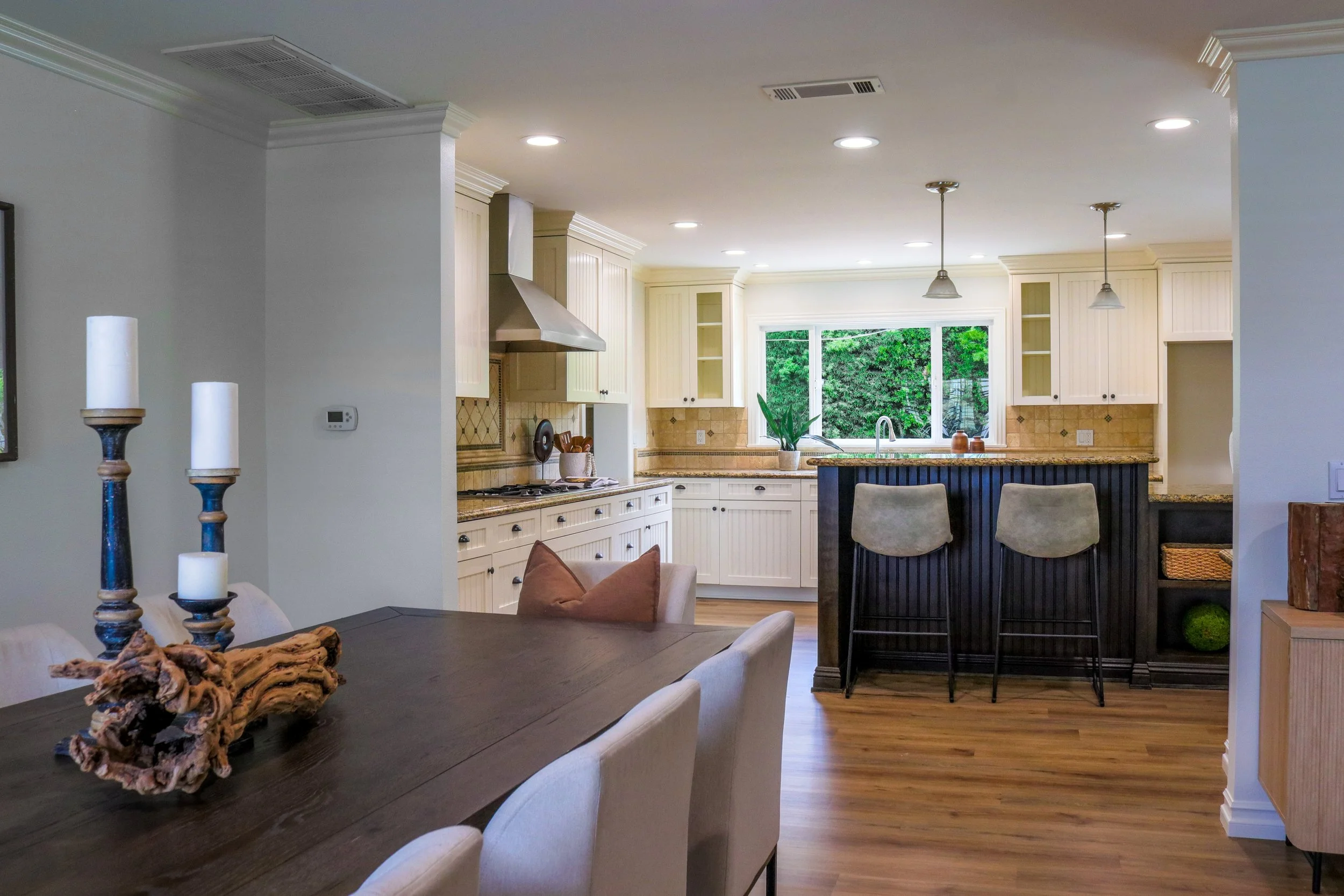 Interior view of a modern kitchen with white cabinets, a window overlooking greenery, a kitchen island with bar stools, and a dining area with a wooden table and candles.