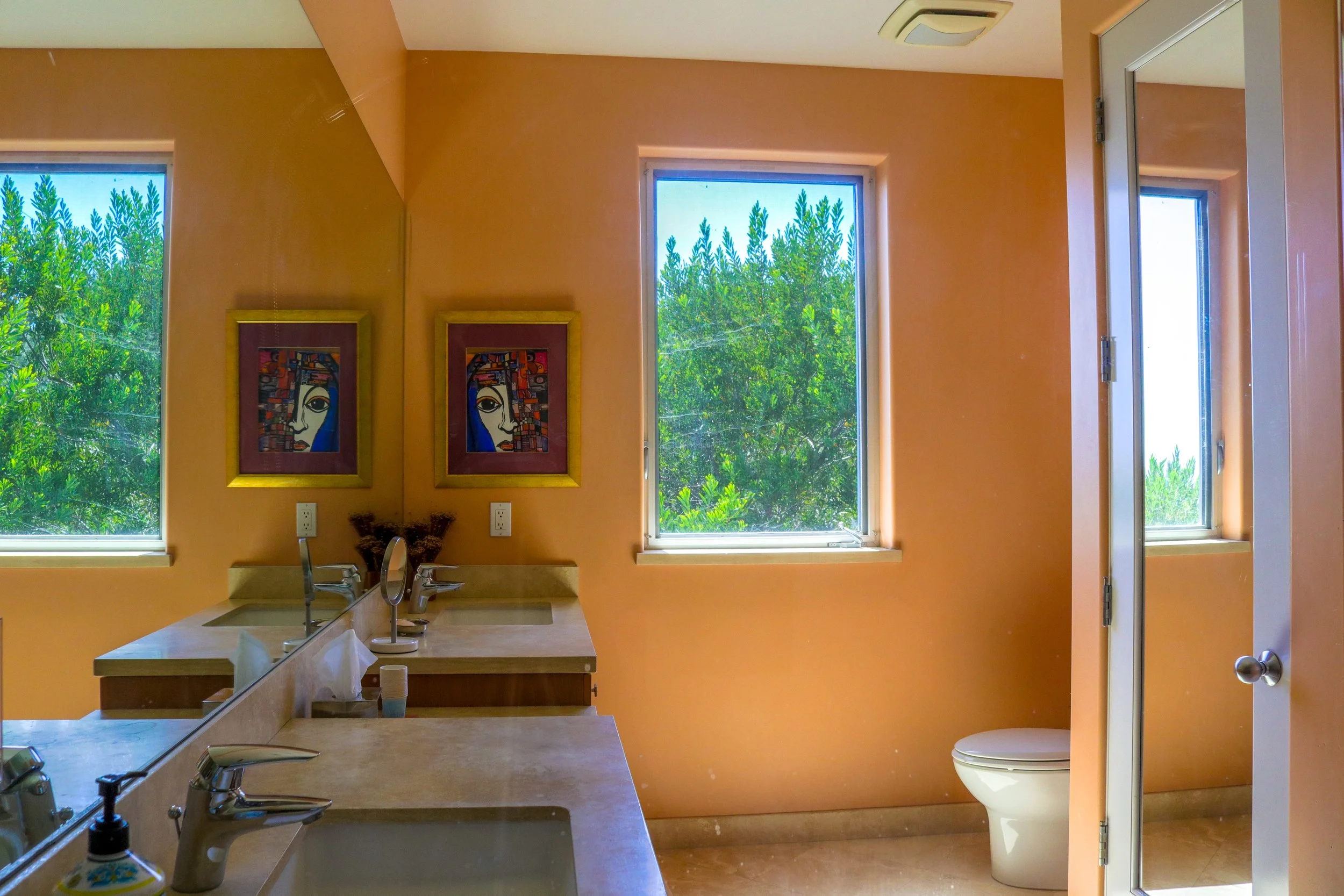 Bathroom with three windows showing greenery outside, orange walls, framed artwork, sinks, and a toilet.