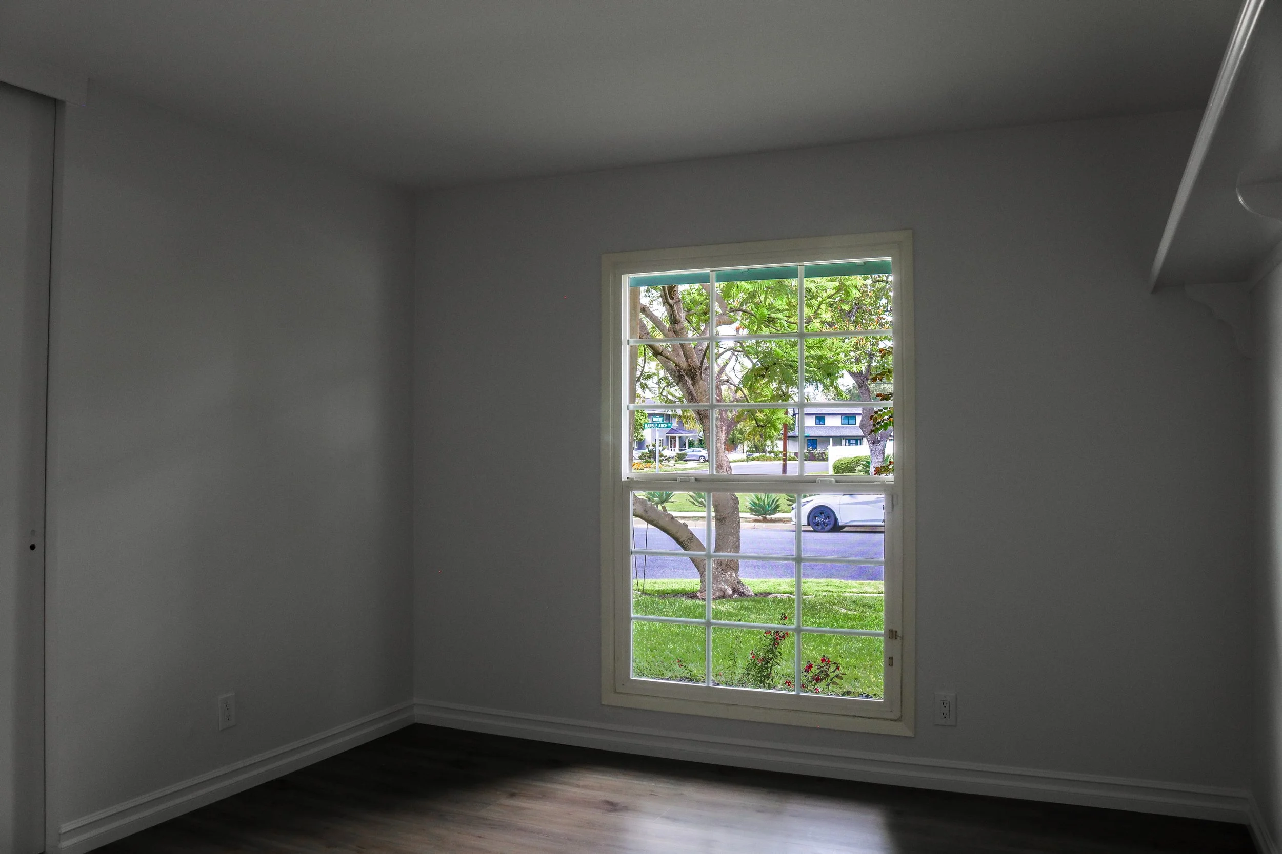 A room with white walls and dark wood flooring, featuring a large window showing trees and a street outside.