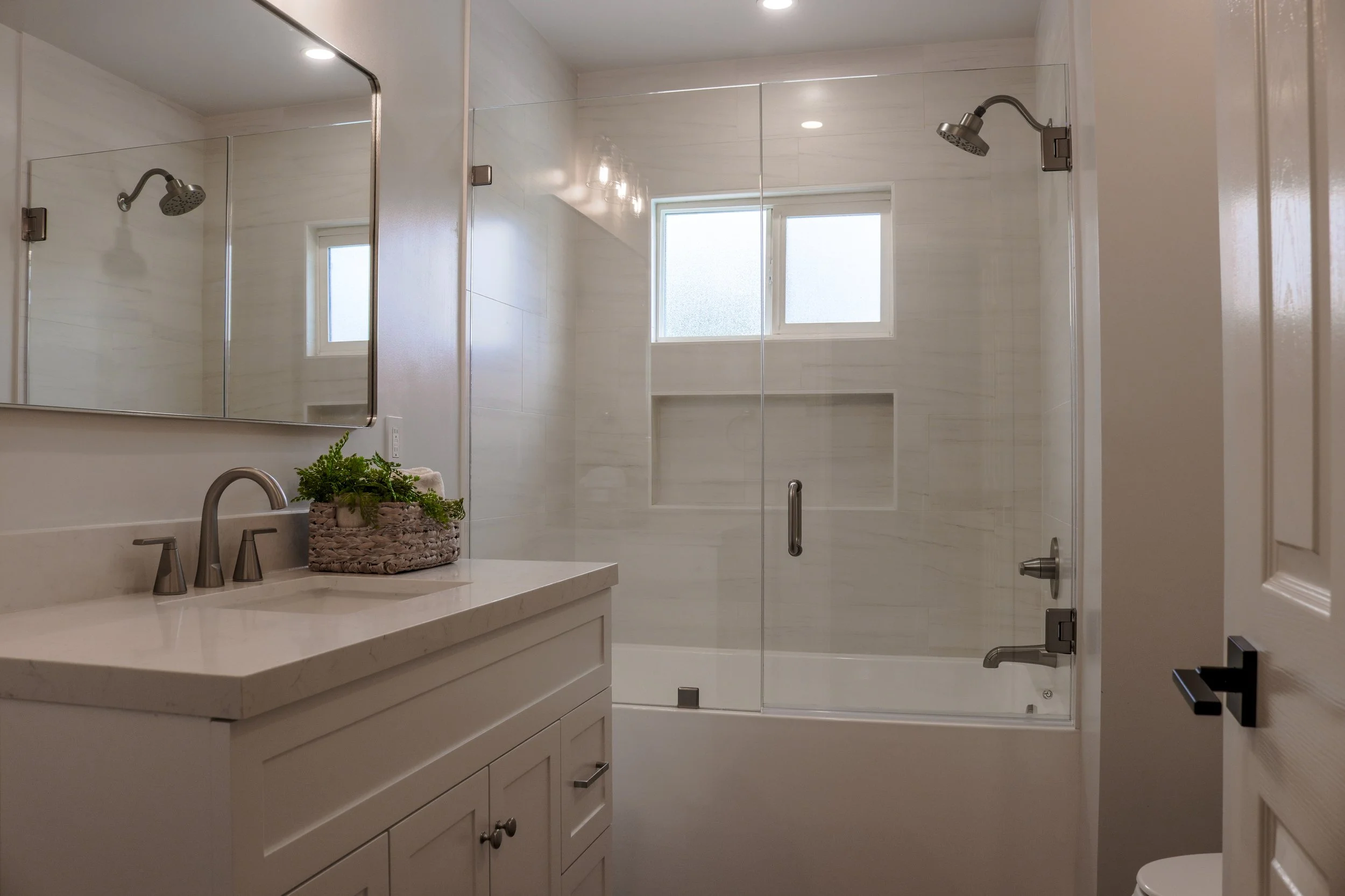 Modern bathroom with a vanity with a marble countertop, a large mirror, a plant, and a glass-enclosed shower with a sliding door, two showerheads, a window, and built-in wall niche.