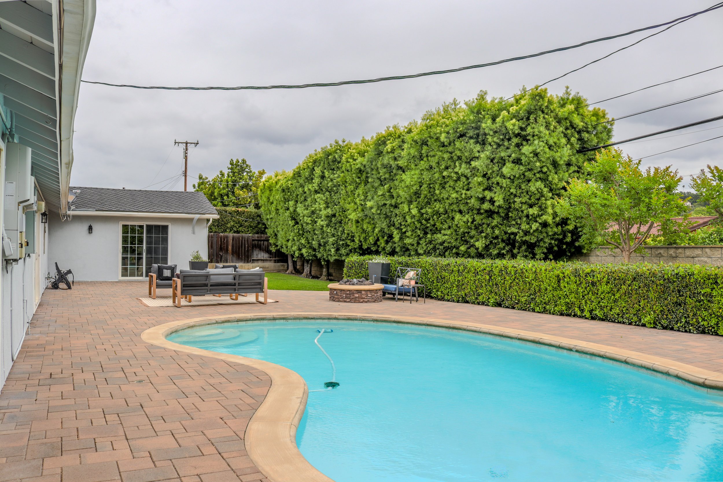 Backyard with a swimming pool, outdoor seating, green shrubs, trees, and overcast sky.