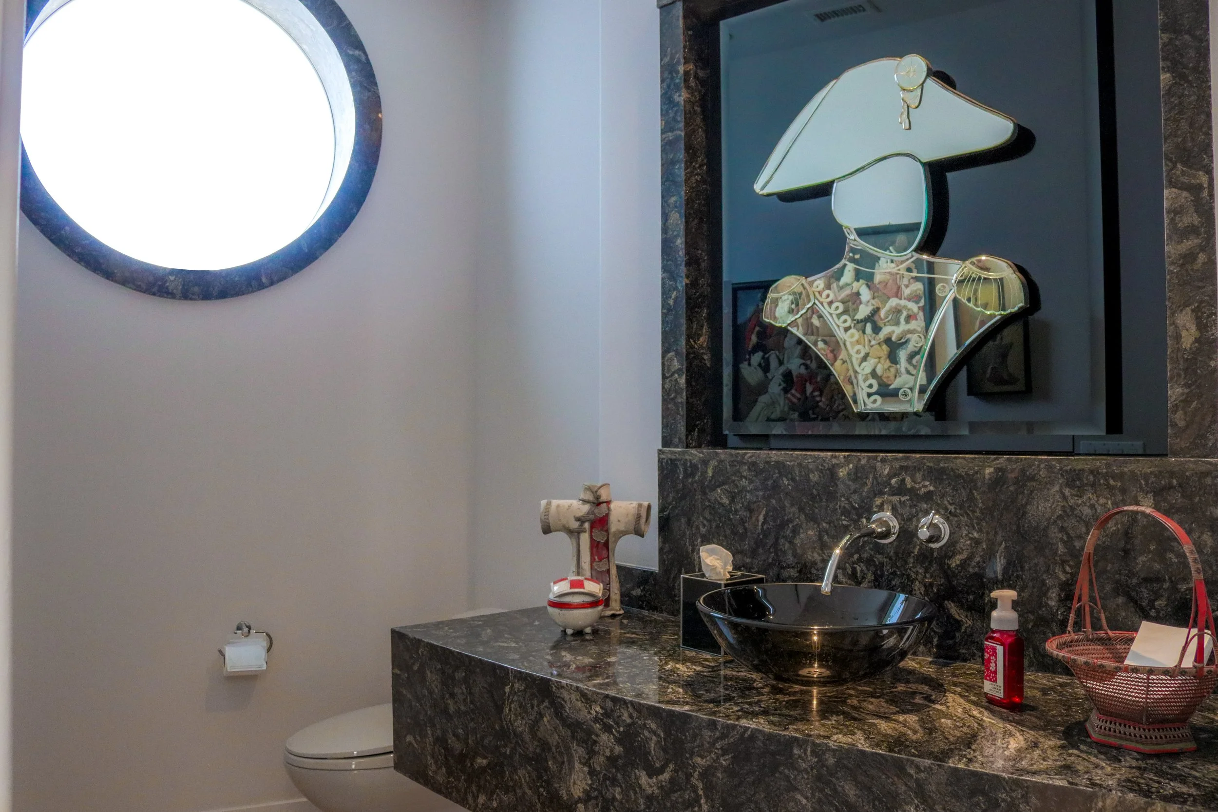 A bathroom counter with a black marble surface, a black vessel sink, and various toiletries, with a mirror and a decorative light fixture above.