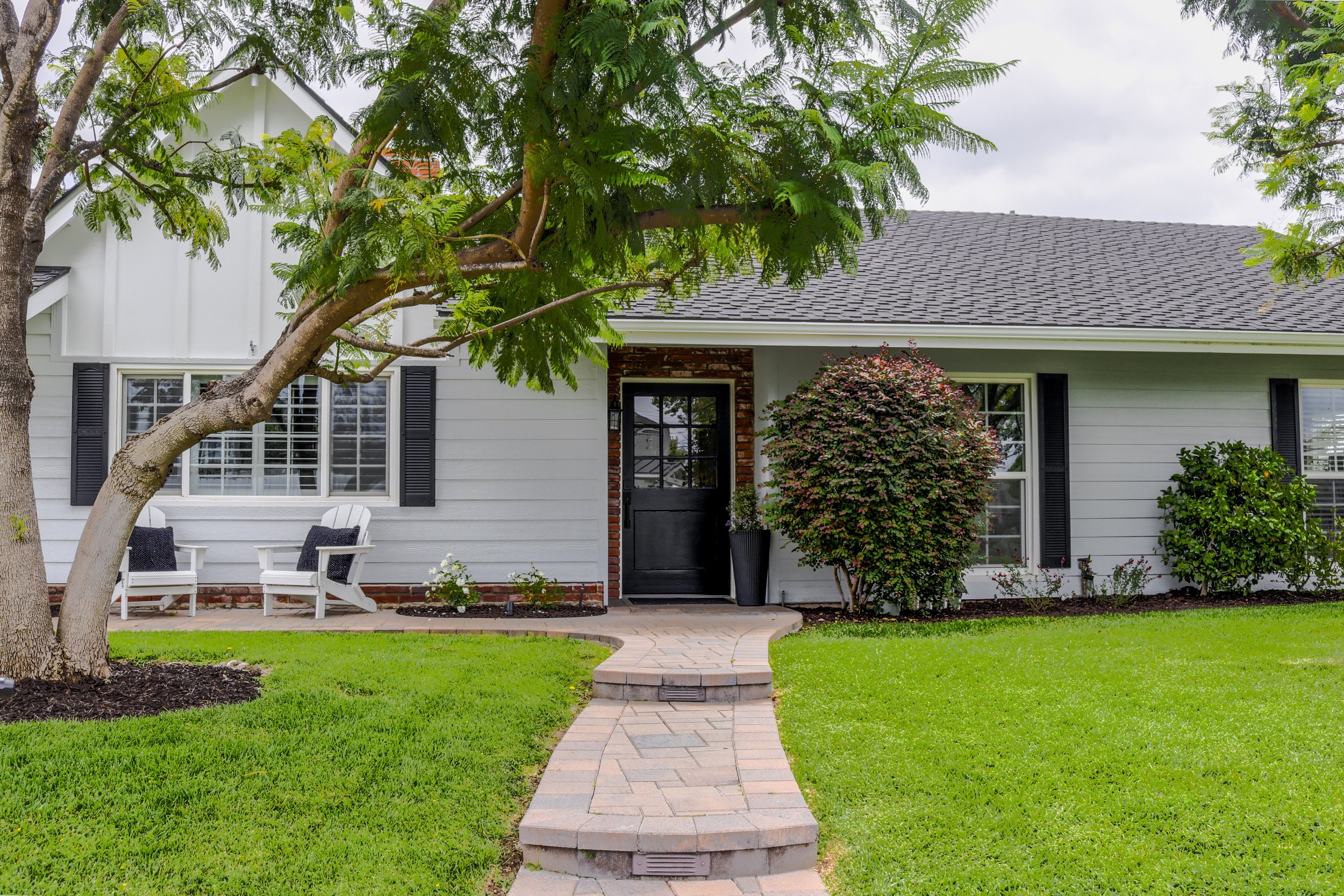 Front yard of a house with a stone pathway, green grass, a large tree, bushes, a black door, and two white chairs.